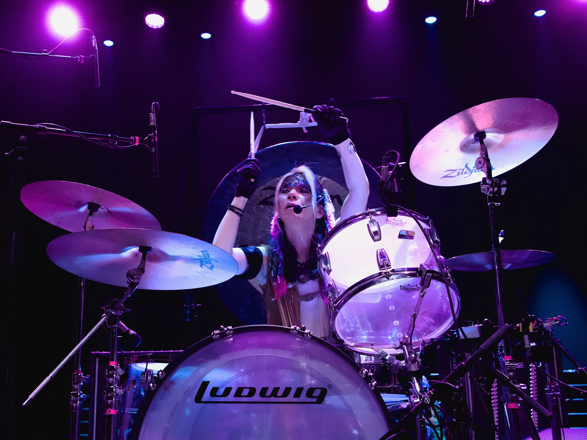 Performer playing a drum set on stage under purple lights, with a Ludwig drum set and cymbals, wearing black gloves, face paint, and colorful hair.