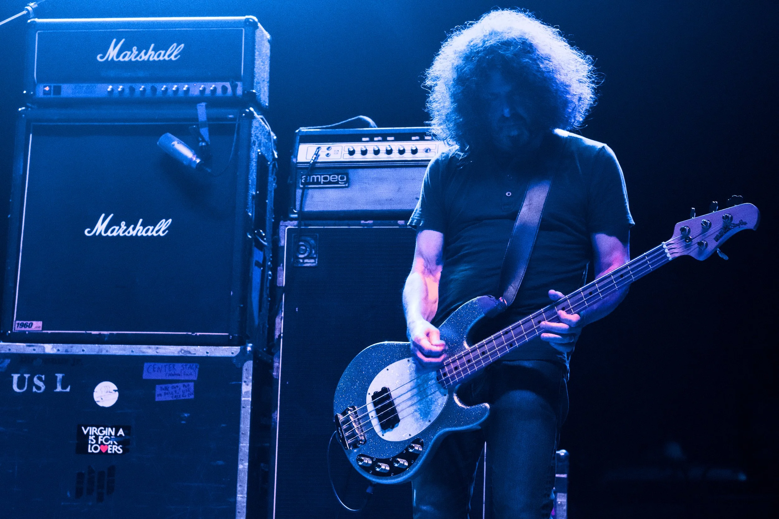 A musician with curly hair playing a bass guitar on stage, surrounded by Marshall amplifiers.