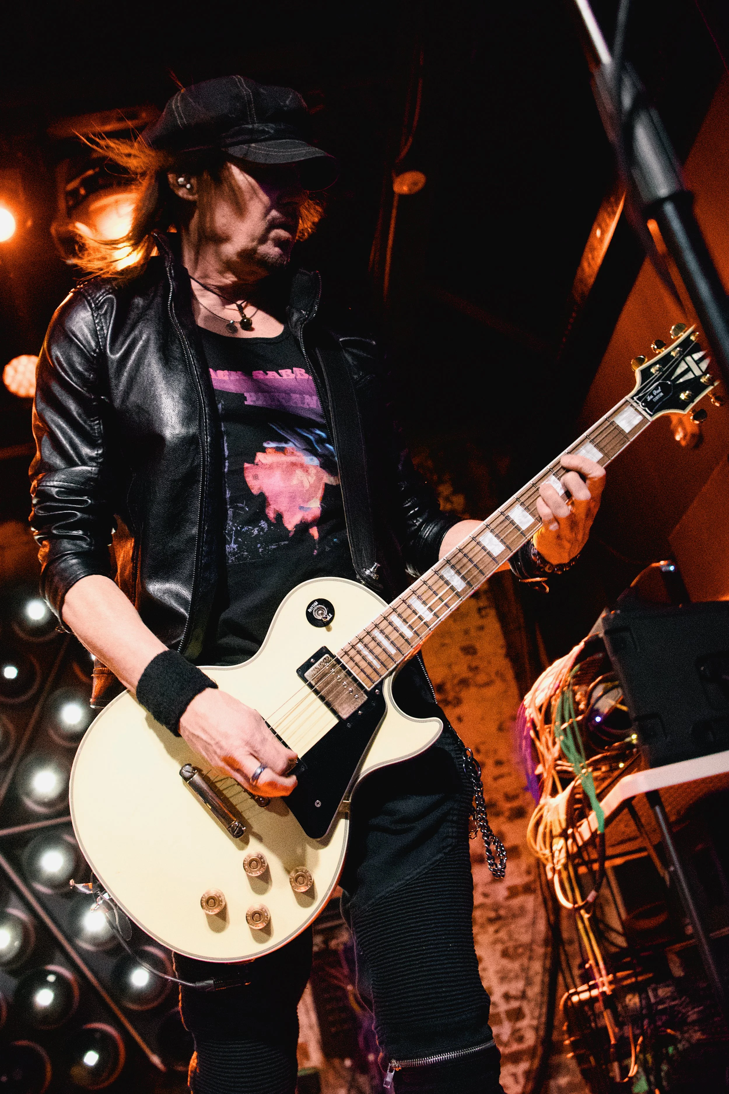 A male guitarist wearing a black leather jacket, black hat, and black clothing performs on stage, playing an electric guitar under orange stage lighting.