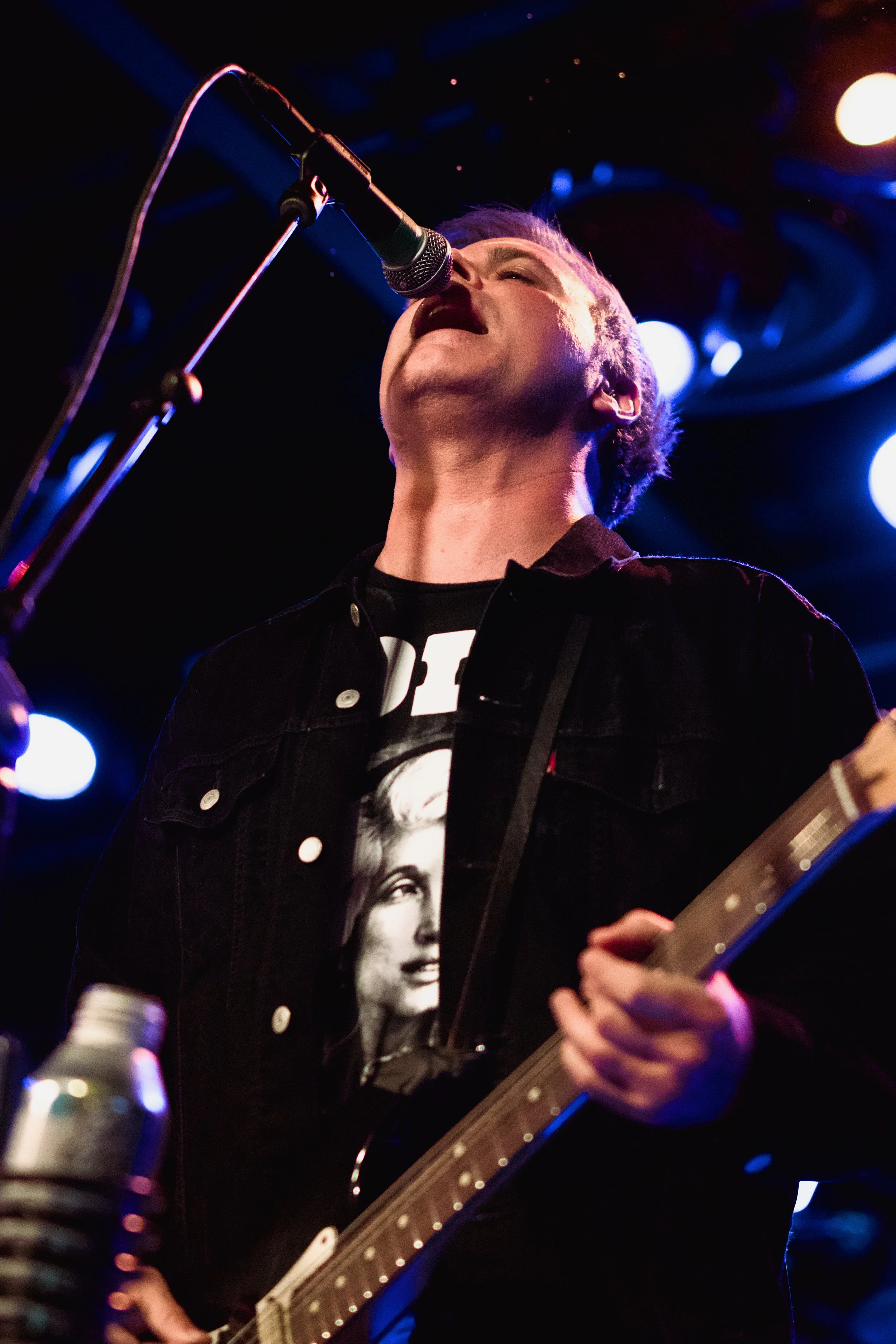 A musician singing into a microphone while playing an electric guitar on stage with colorful lights.
