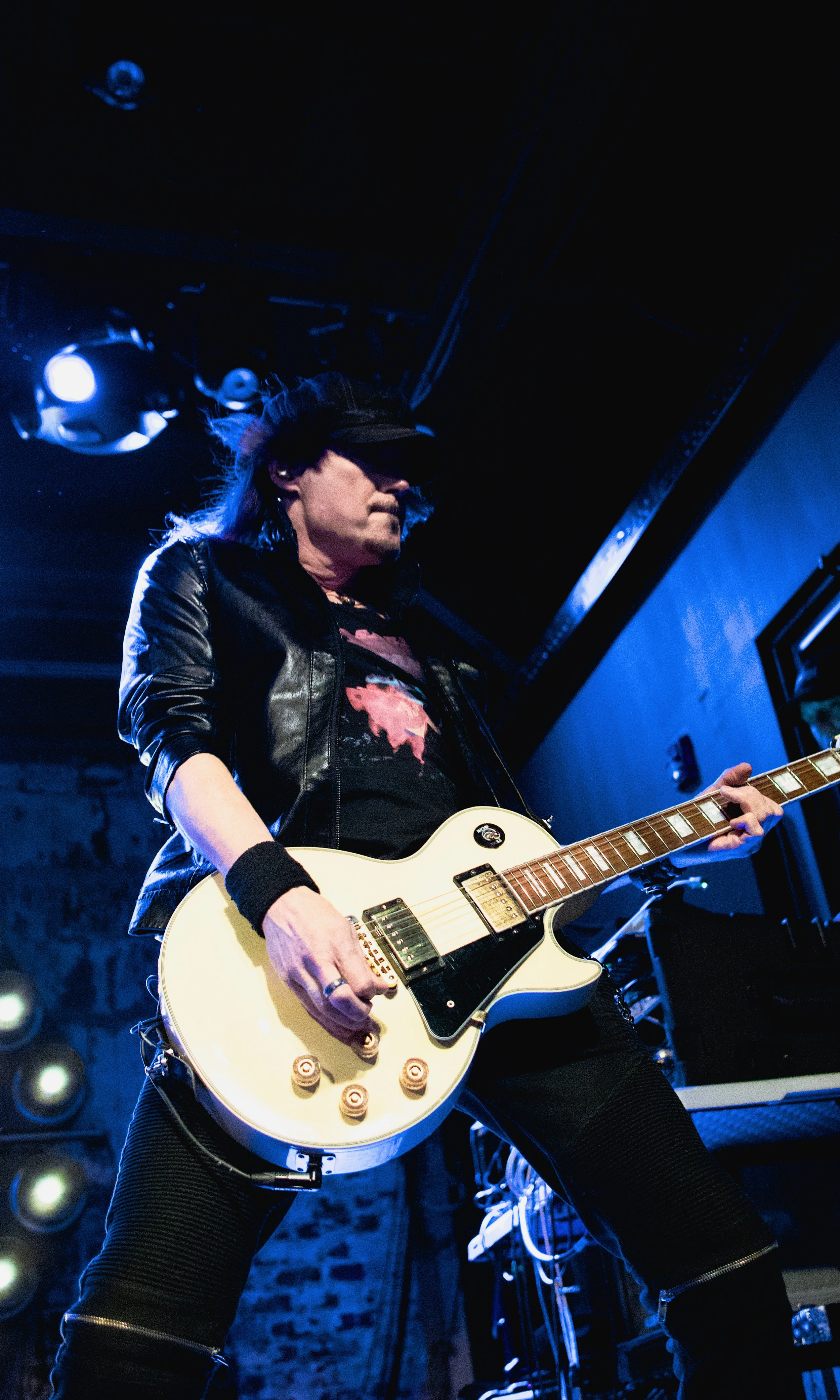 A guitarist in a black leather jacket, dark cap, and sunglasses performing on stage with blue stage lighting.