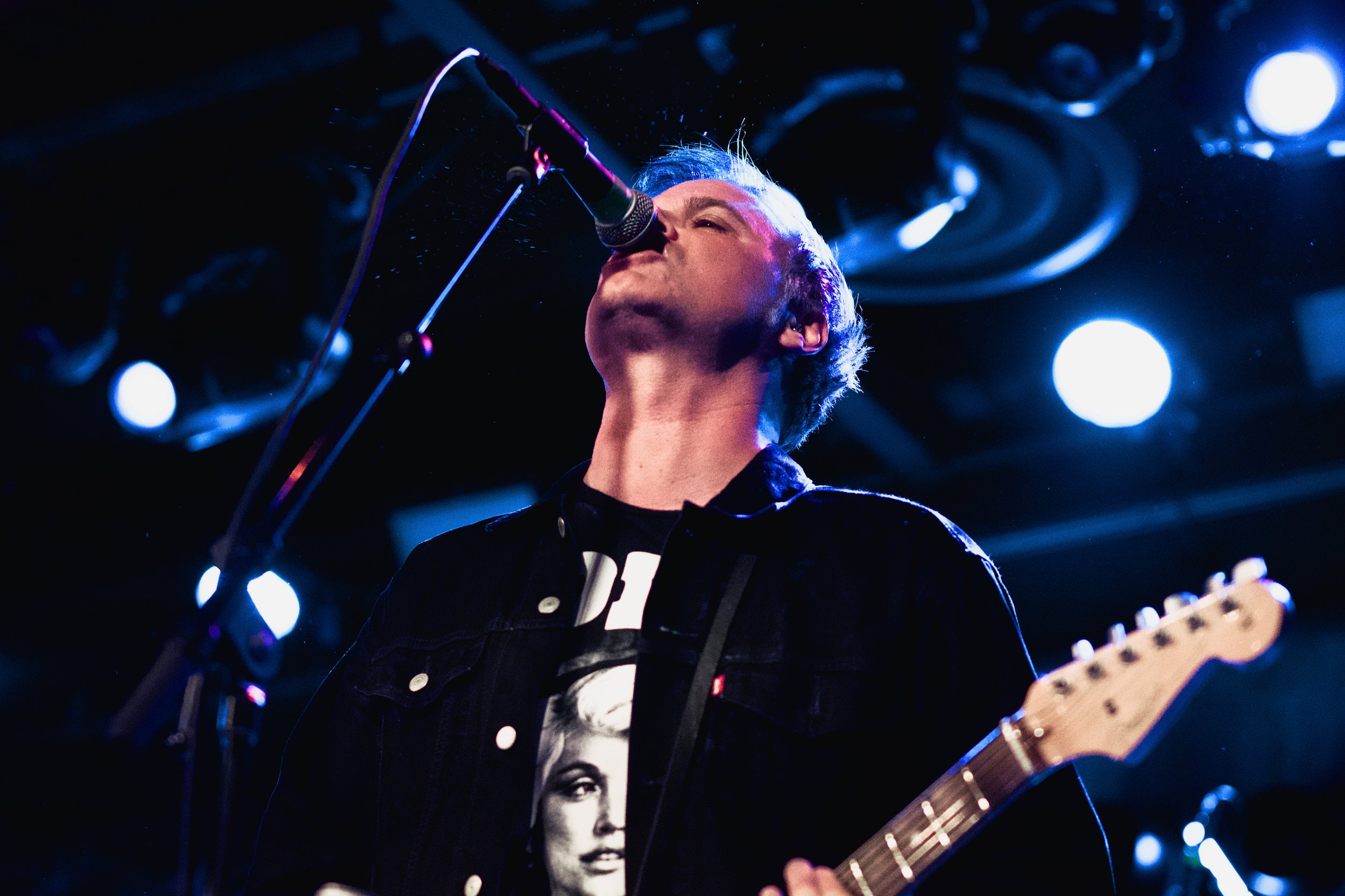 A musician singing into a microphone while playing an electric guitar on a stage with blue lighting.