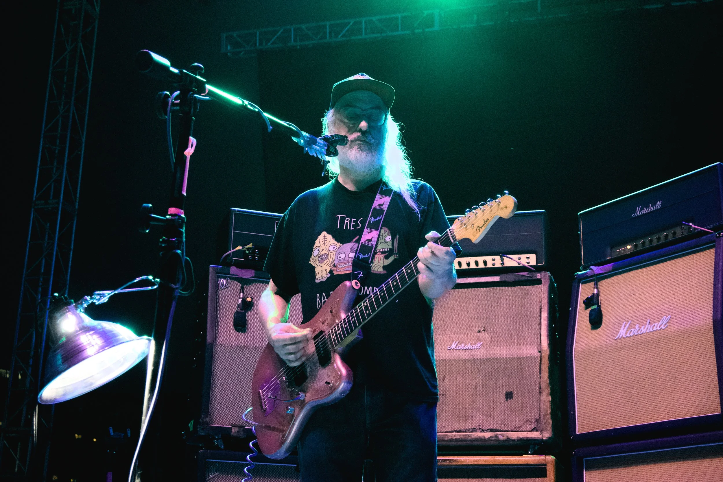 A man with long gray hair and a beard playing an electric guitar on stage with Marshall amplifiers behind him, illuminated by green and blue stage lighting.