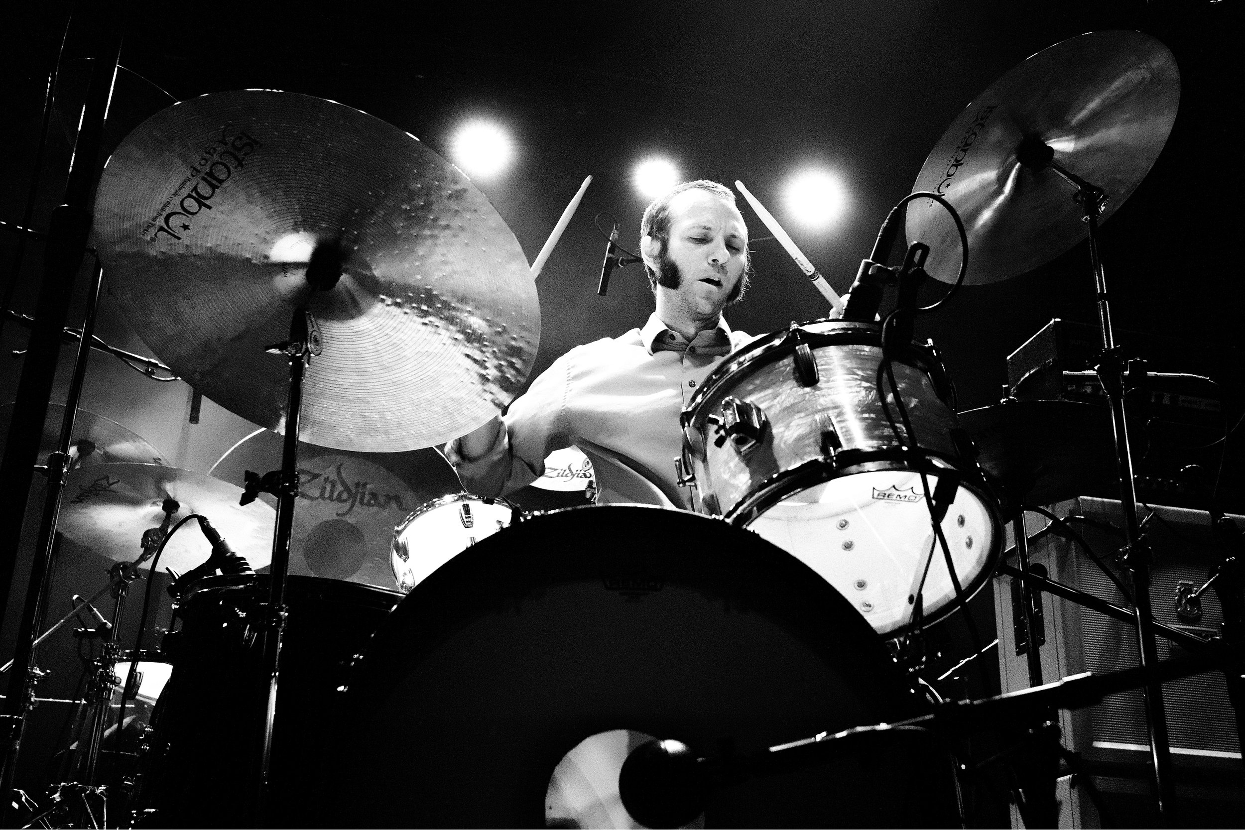 Black and white photo of a man playing the drums on stage, with a focused expression, illuminated by stage lights.