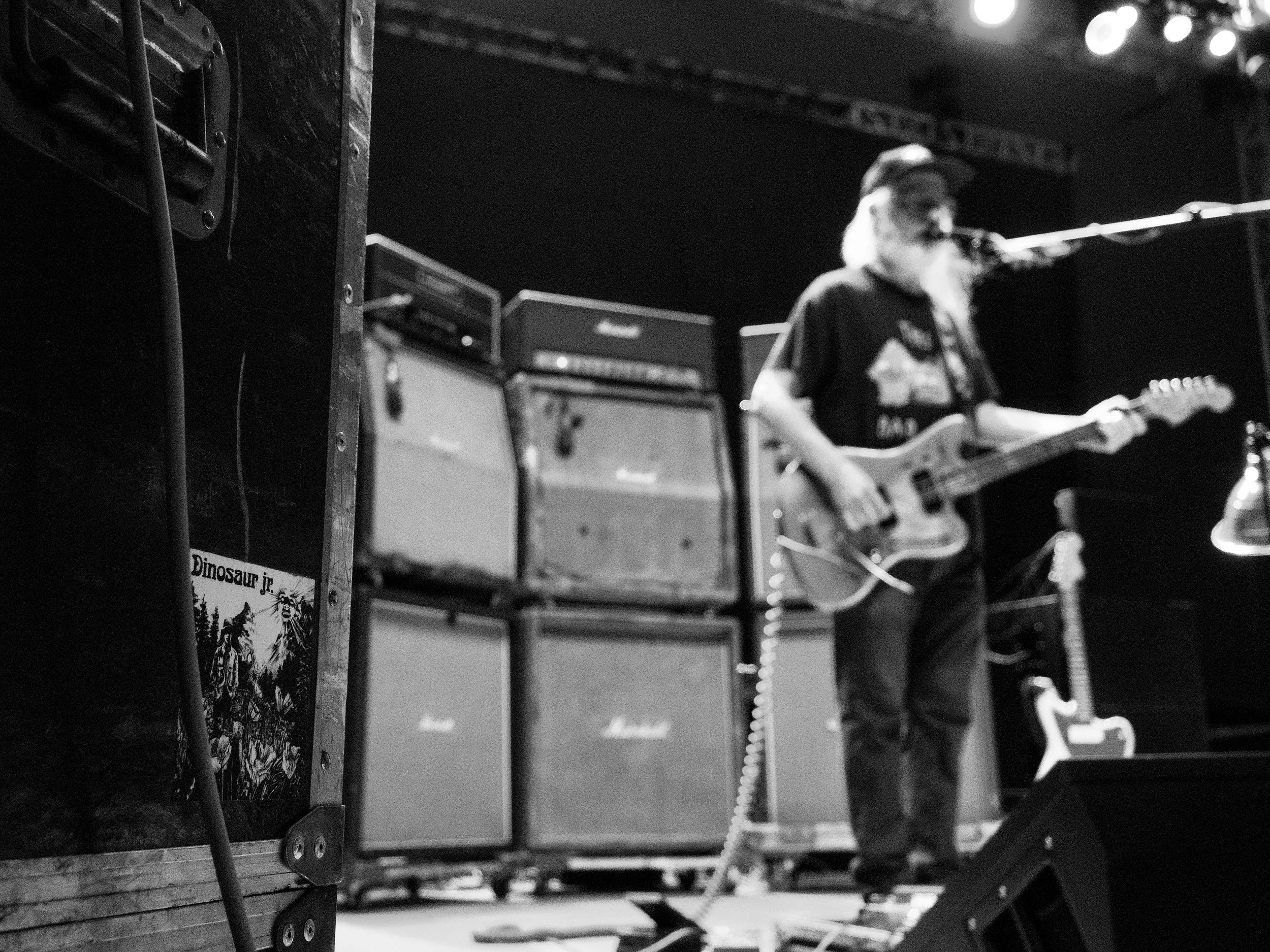 A musician with a beard and glasses playing an electric guitar on stage, with multiple bass amplifiers behind him. The scene appears to be a live performance or rehearsal.