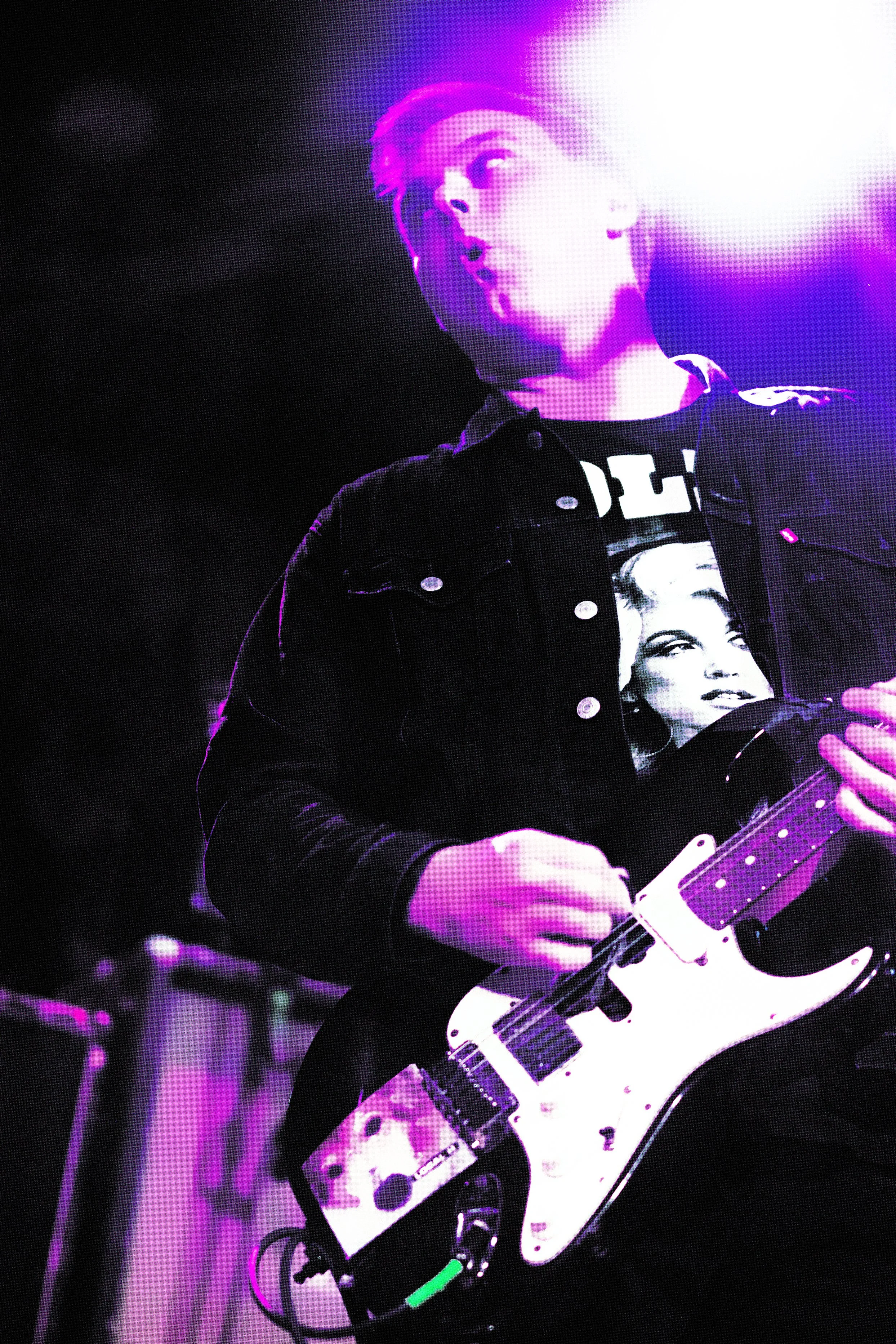 Young man playing an electric guitar on stage with purple and white lighting.