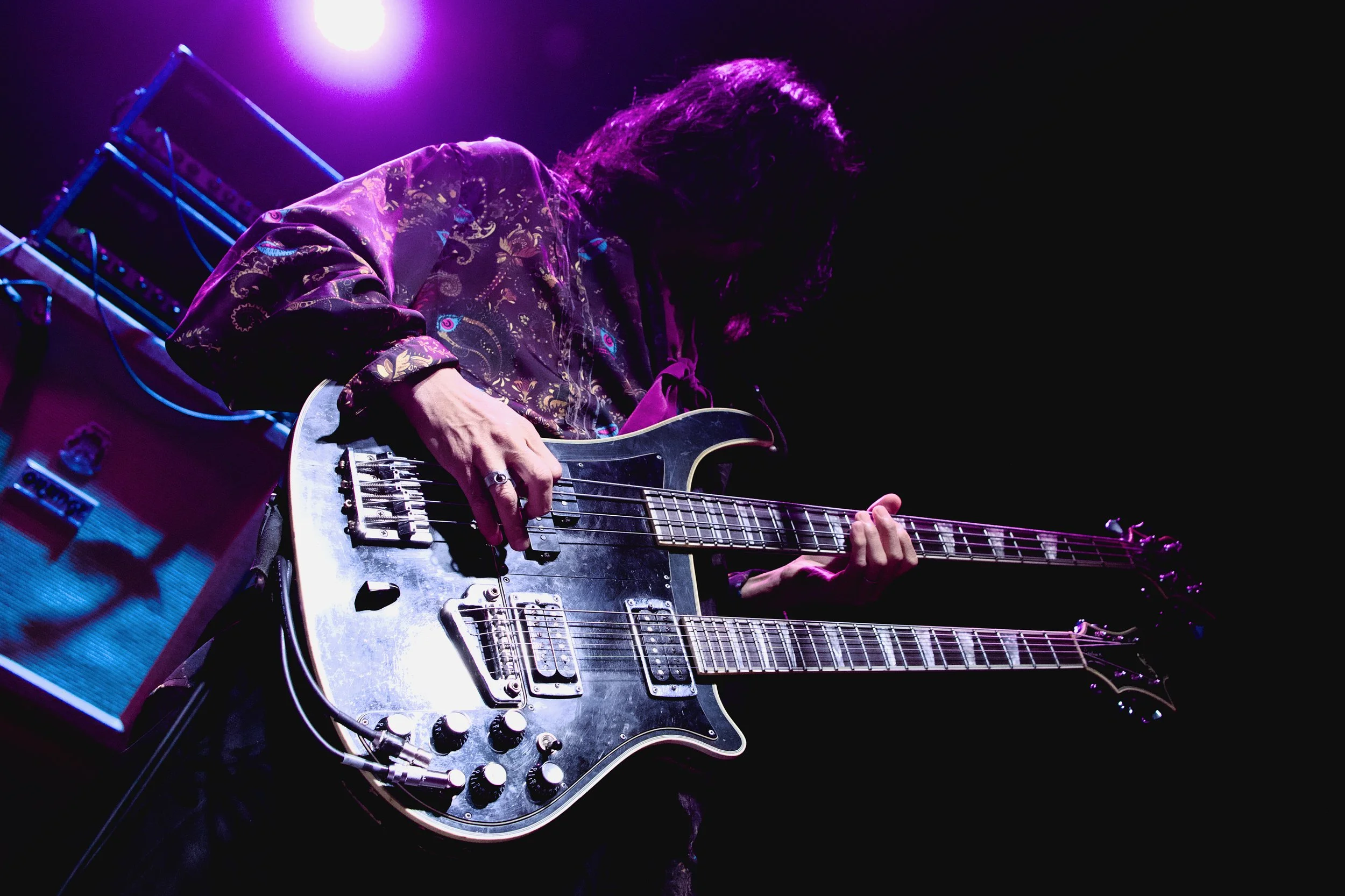 Musician playing a black double-neck electric guitar on stage, illuminated by purple stage lights, with amplifier in the background.