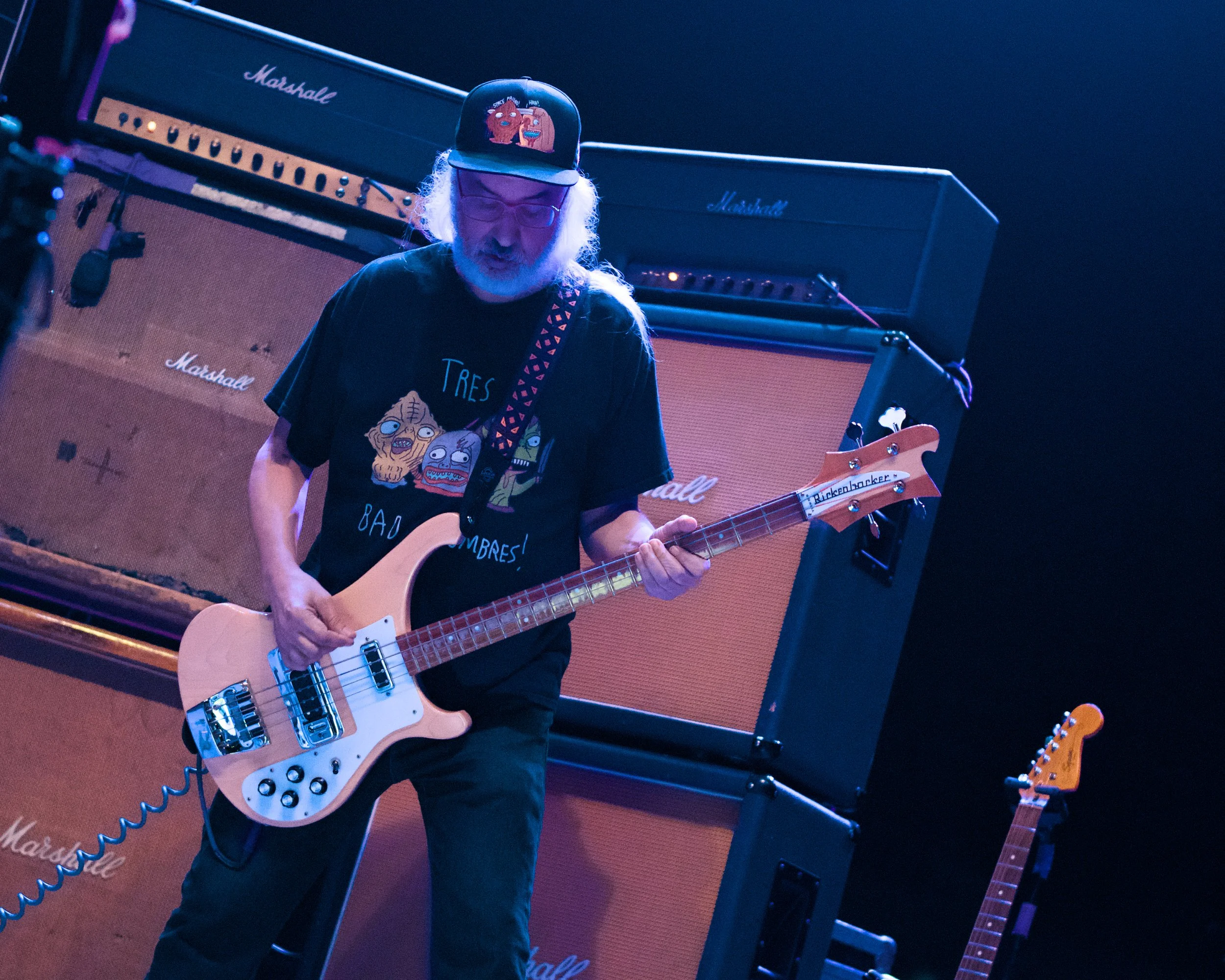 A man with gray hair and glasses playing a cream-colored bass guitar on stage, in front of large orange Marshall amplifiers.