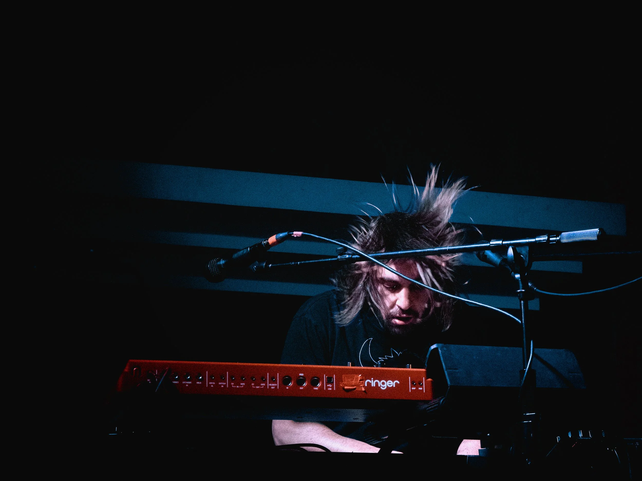 A musician with spiky hair playing a red keyboard on stage with a microphone overhead, dim lighting and dark background.