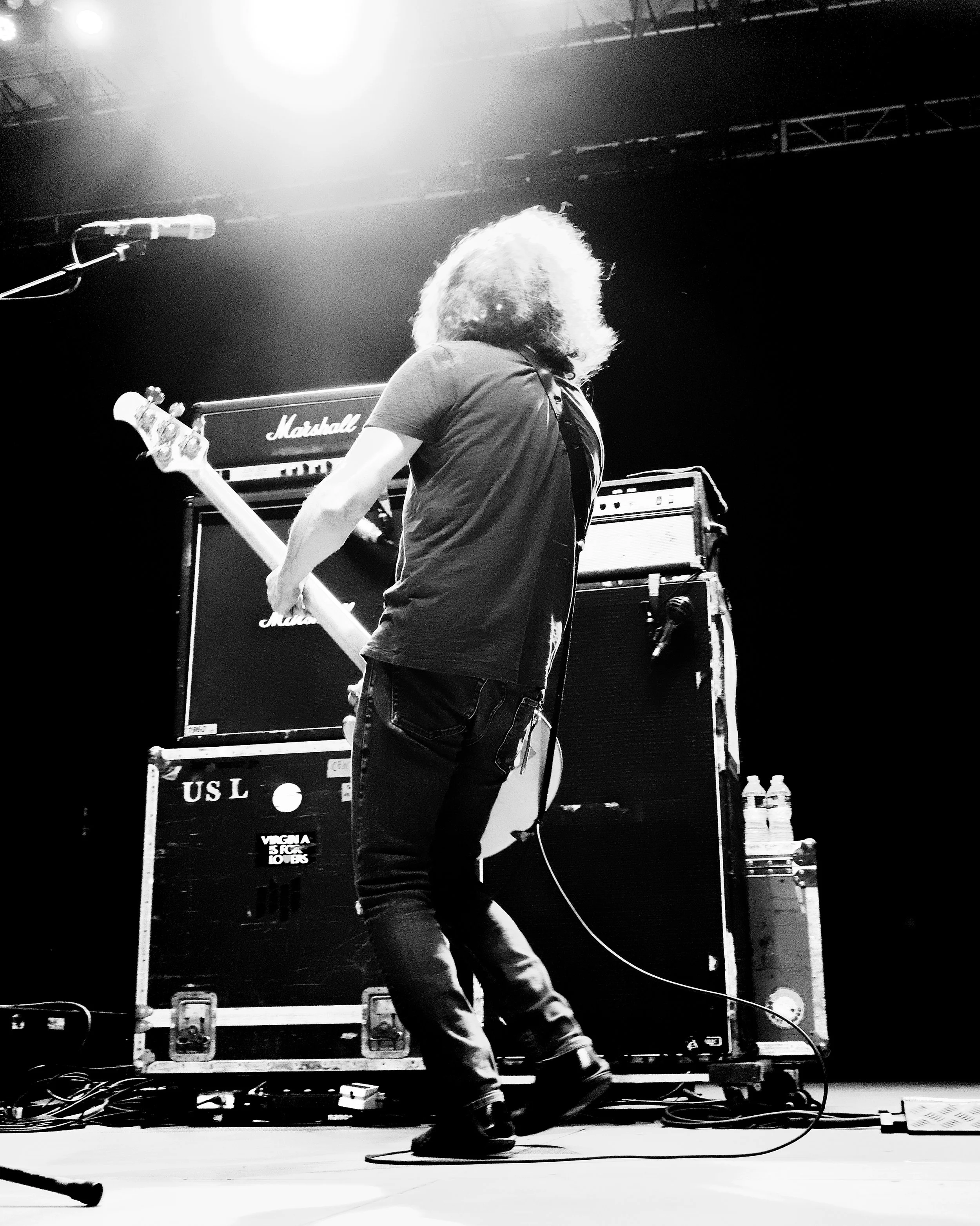 A musician with long hair playing an electric bass guitar on stage behind an amplifier, with stage lights overhead.
