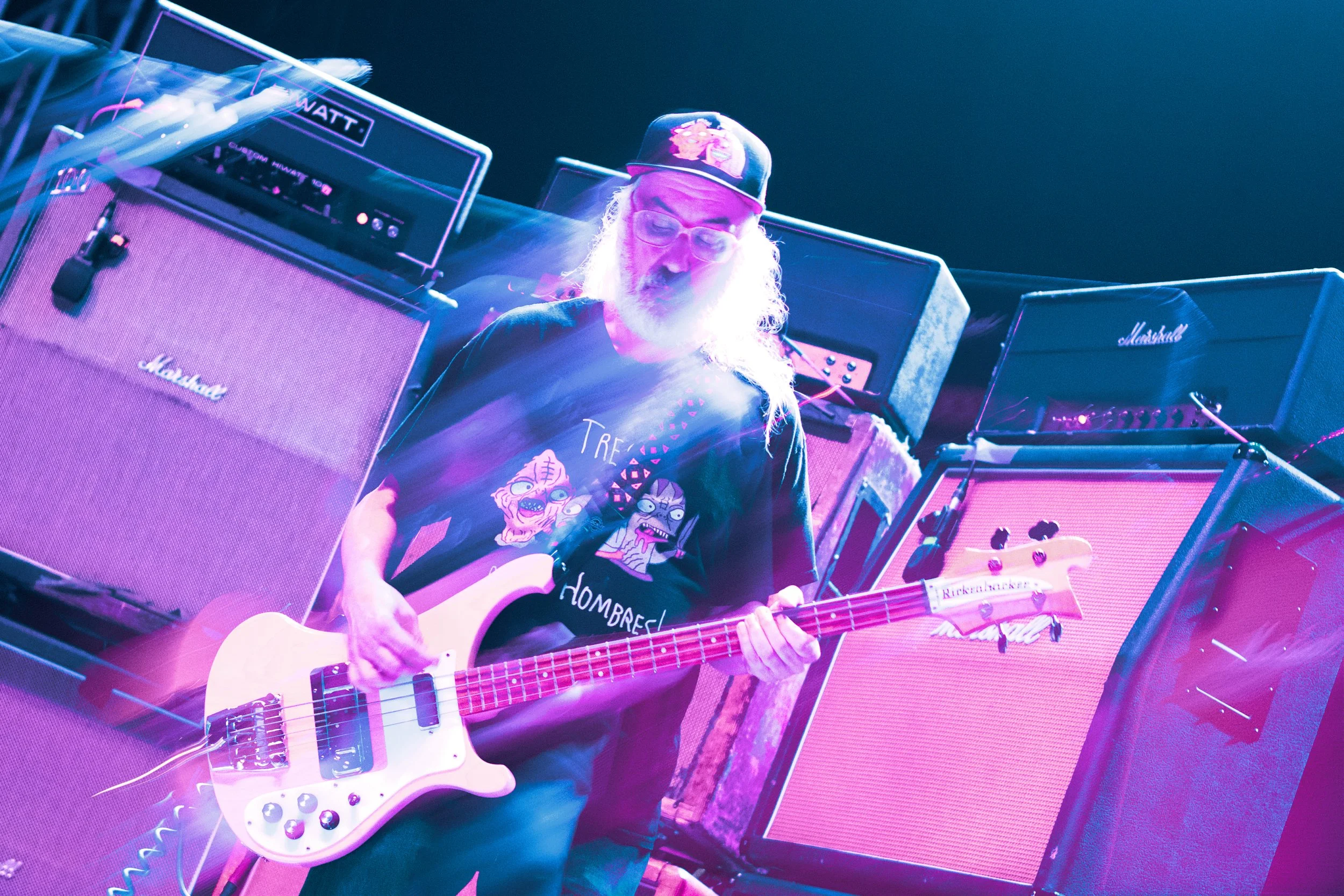 A bearded man wearing glasses and a colorful cap playing an electric bass guitar surrounded by multiple large guitar amplifiers in a music performance setting with colorful stage lighting.
