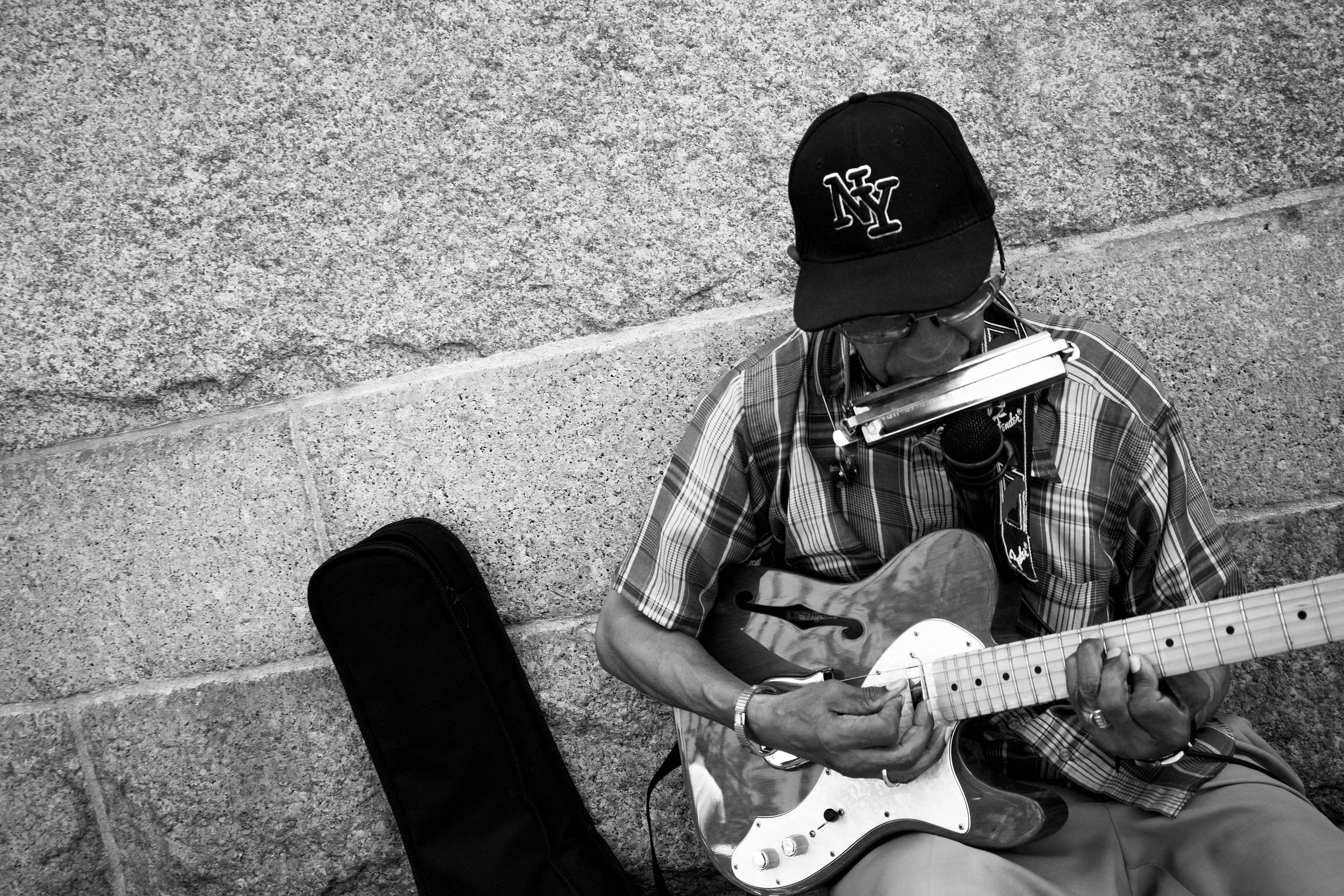 A man wearing a baseball cap and plaid shirt playing an electric guitar on the street, with a harmonica holder around his neck and a harmonica attached, sitting against a concrete wall.