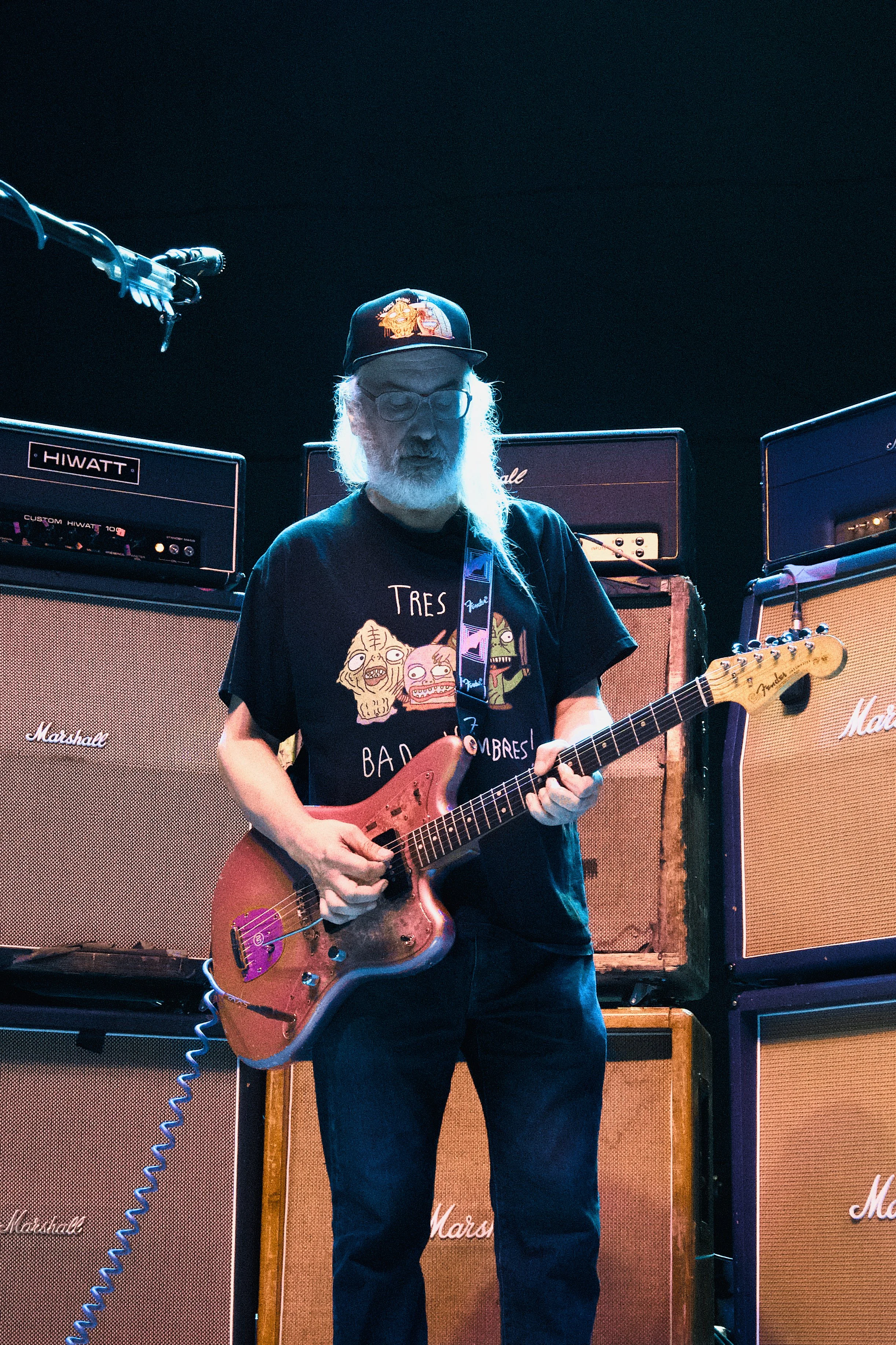 An older man with glasses and a beard playing an electric guitar on stage, surrounded by Marshall amplifier stacks.