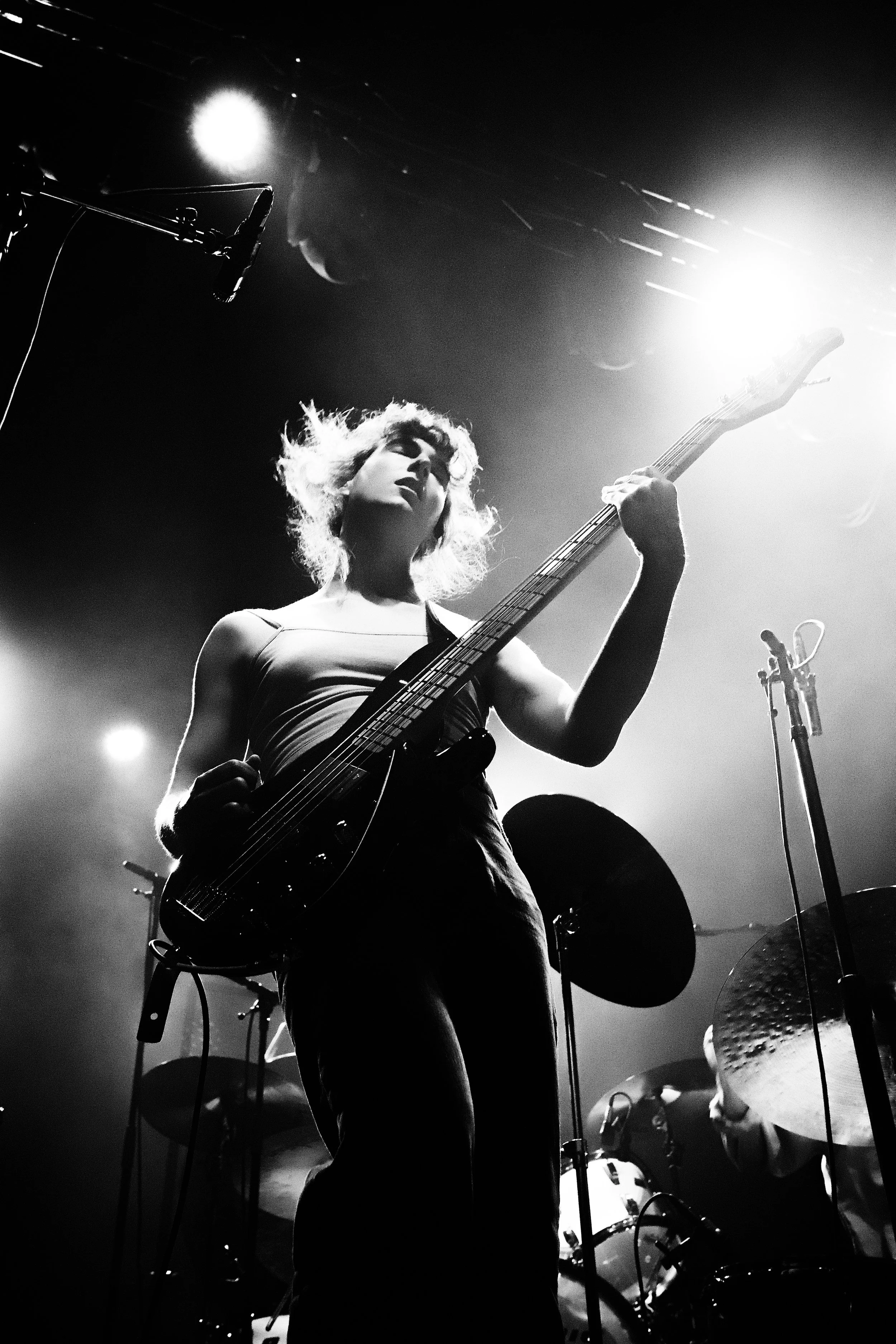 A black and white photo of a female guitarist playing on stage, with a drummer visible in the background. The stage lighting creates a bright halo around her head.
