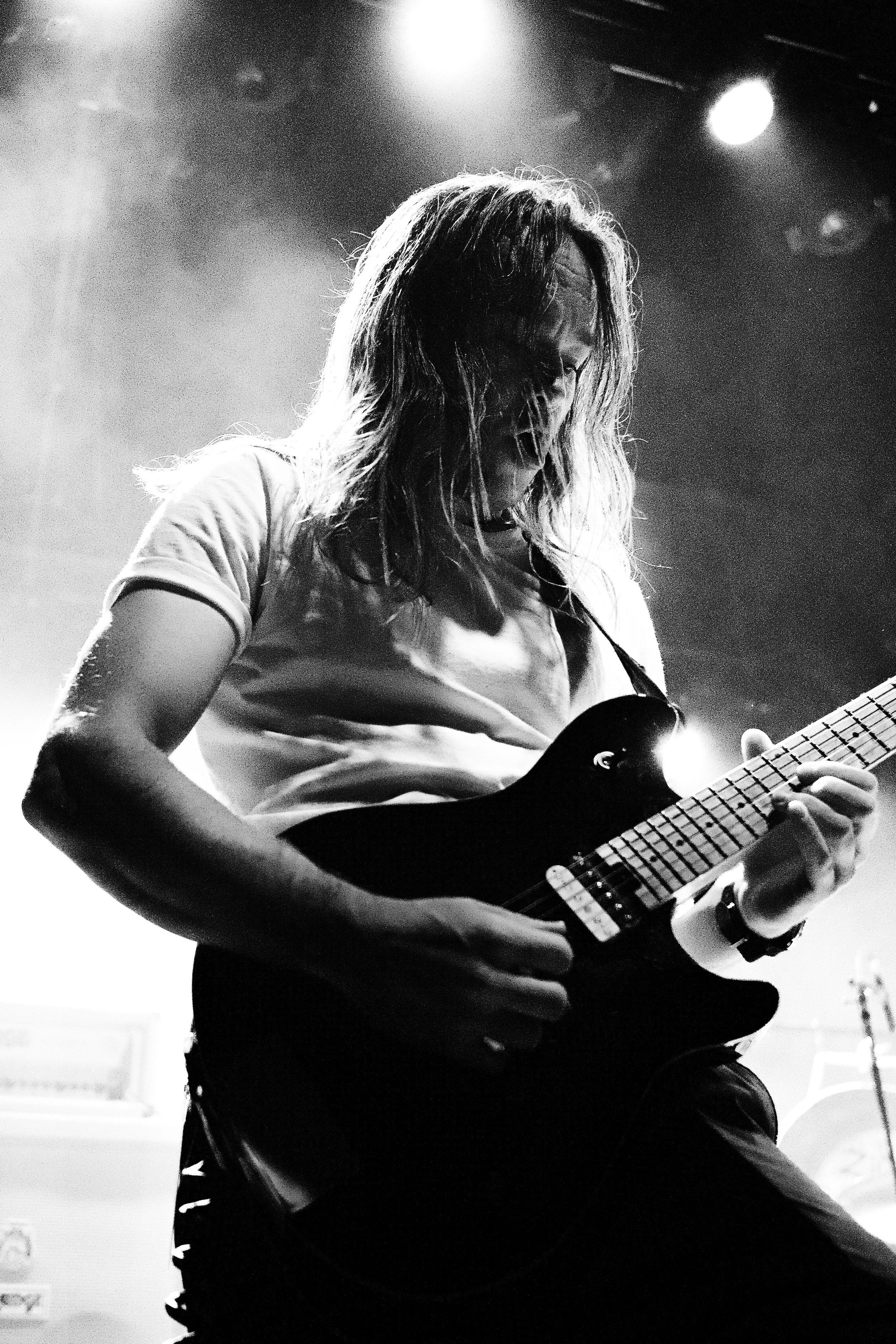 A black and white photo of a musician with long hair playing an electric guitar on stage, with stage lights shining down.