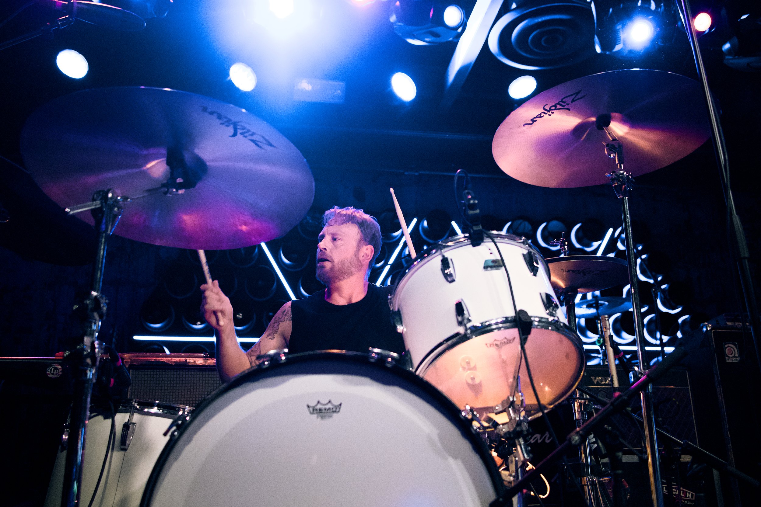 A male drummer with a beard and tattoos on his arm playing a white drum set on stage illuminated by blue and purple stage lights.