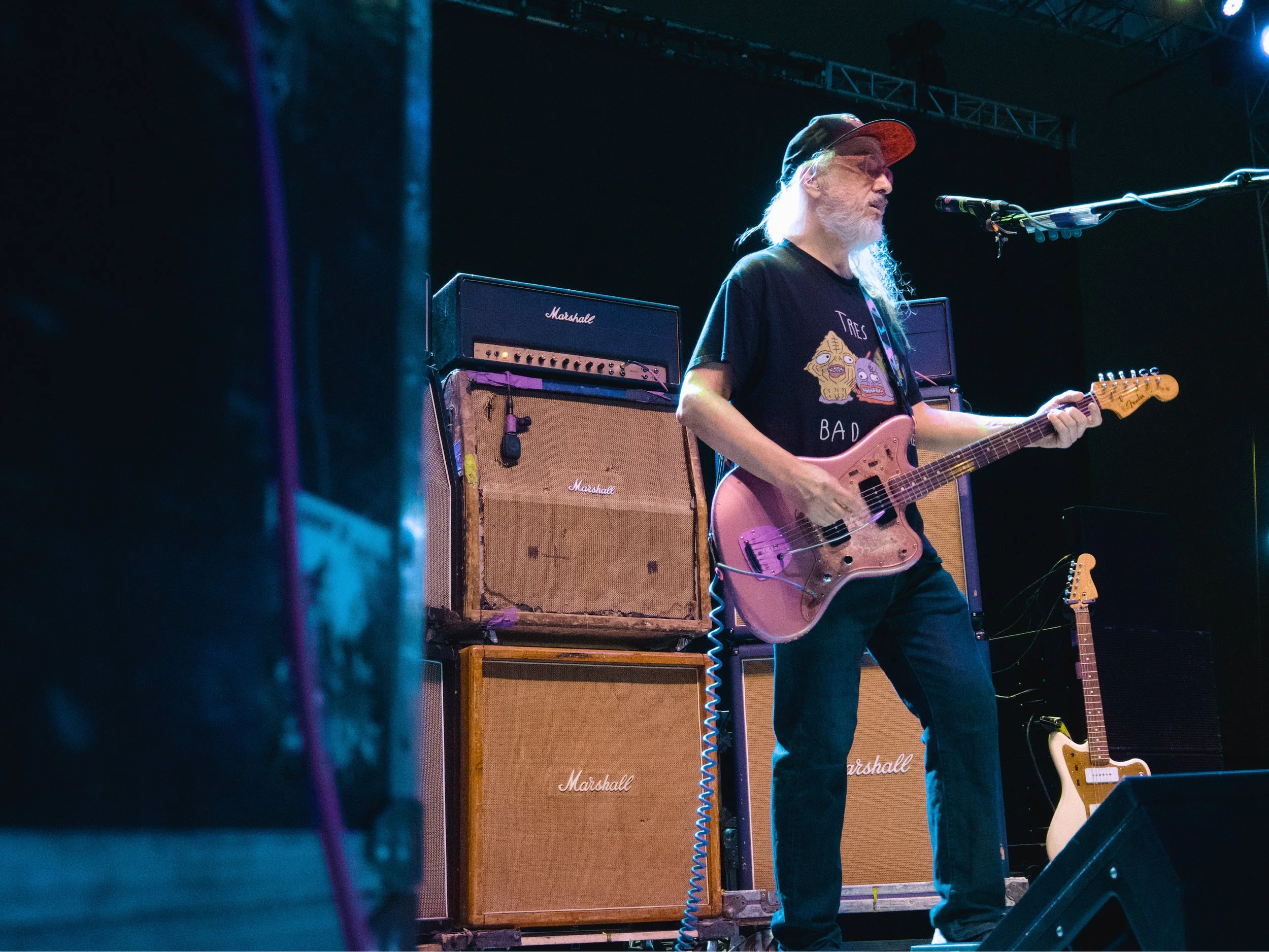 A musician with long gray hair, a beard, and glasses wearing a black T-shirt with colorful graphics, playing a guitar on stage with amplifiers behind him.