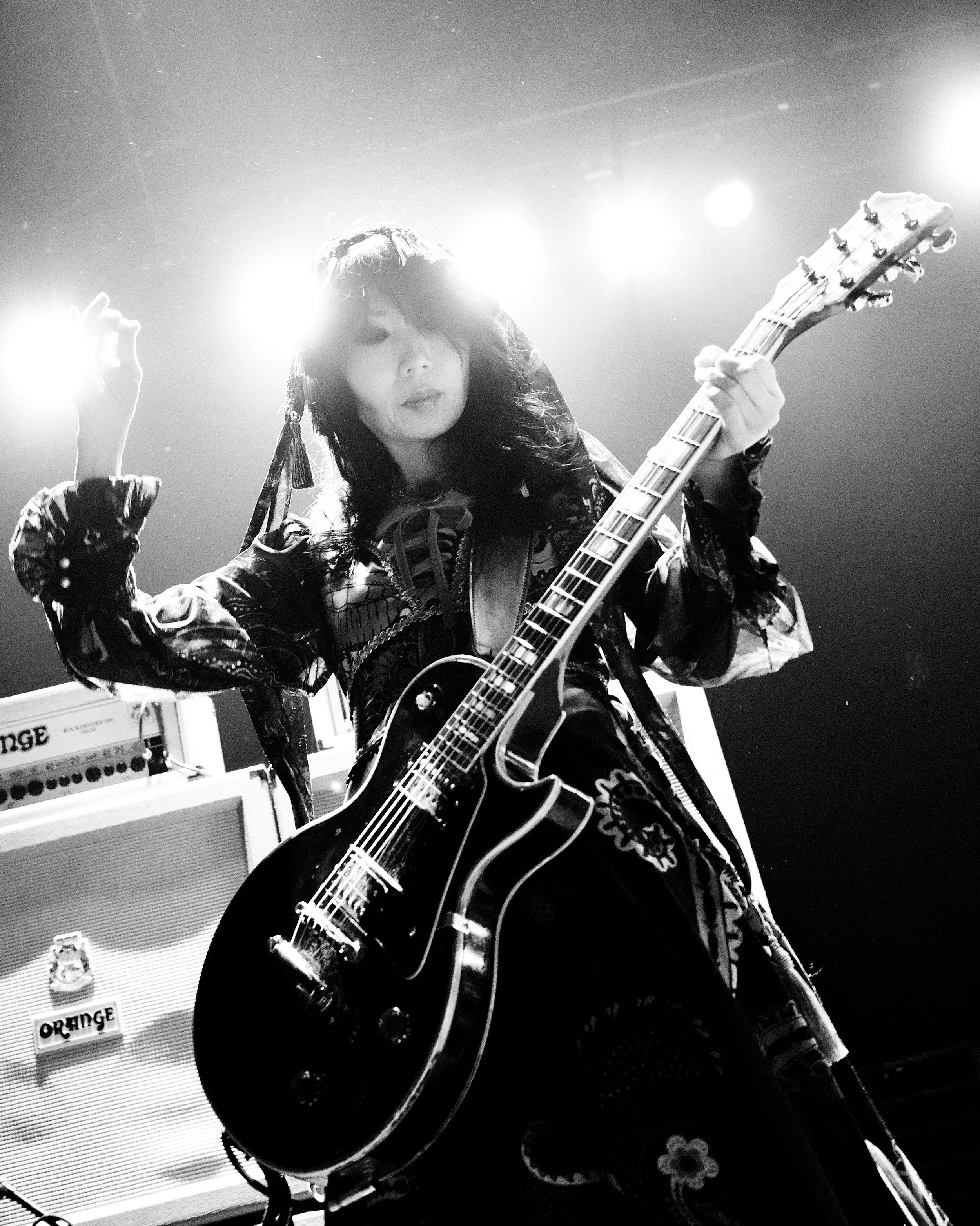A female guitarist performing on stage with bright lights behind her, playing an electric guitar.