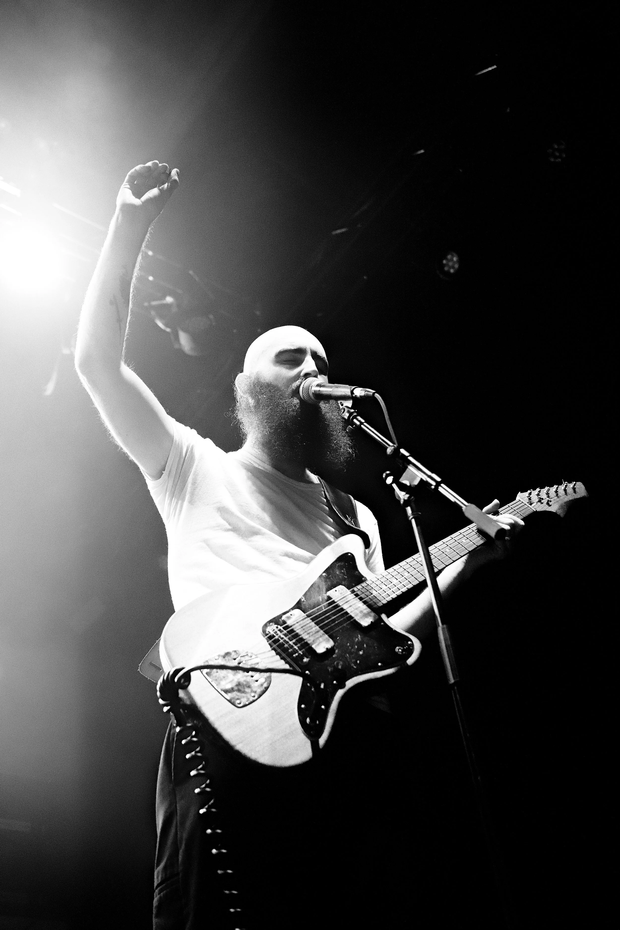 Black and white photo of a bearded male musician singing into a microphone while playing an electric guitar on stage, with one arm raised.