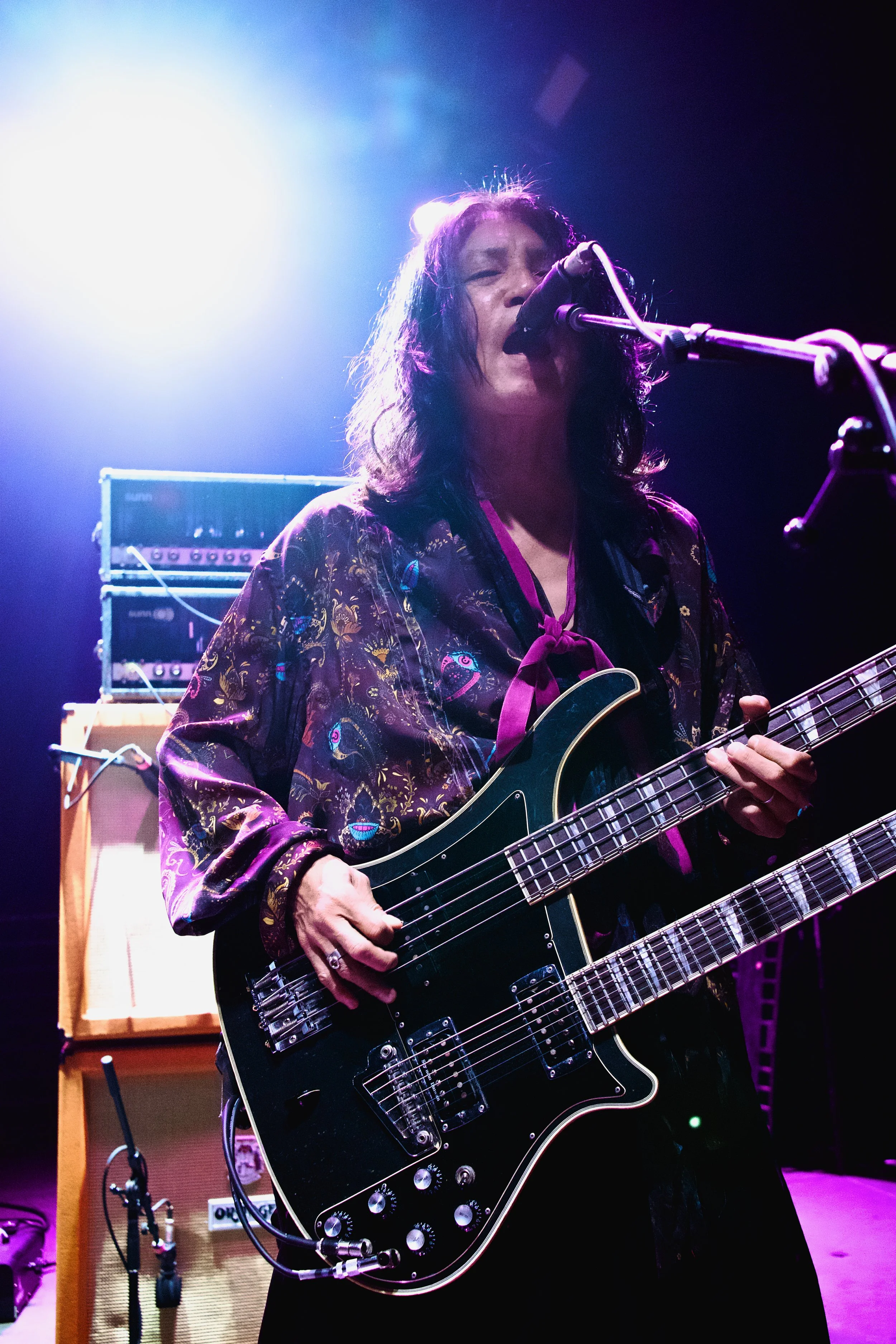 A woman singing and playing a black electric guitar on stage, with purple and blue lighting and guitar amplifiers in the background.
