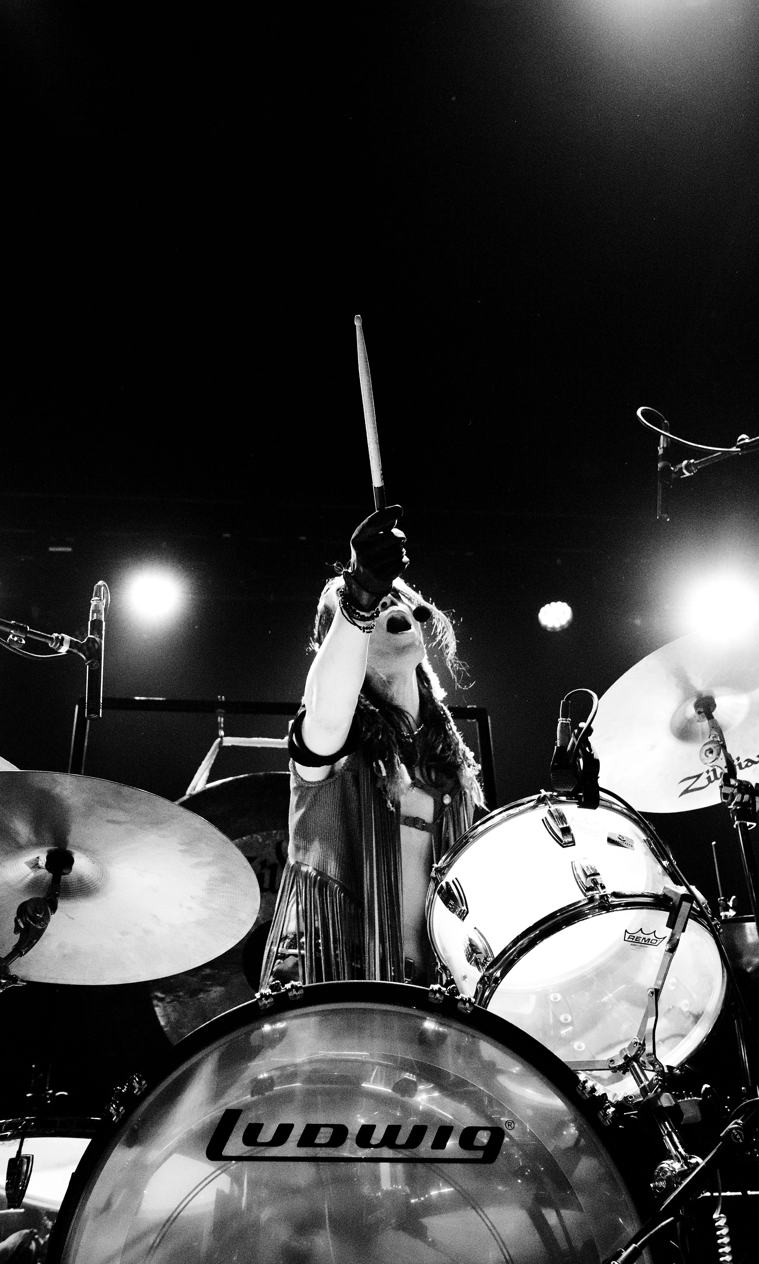 A female drummer on stage during a live performance, raising her drumstick in the air, surrounded by drums and cymbals, with stage lights shining behind her.