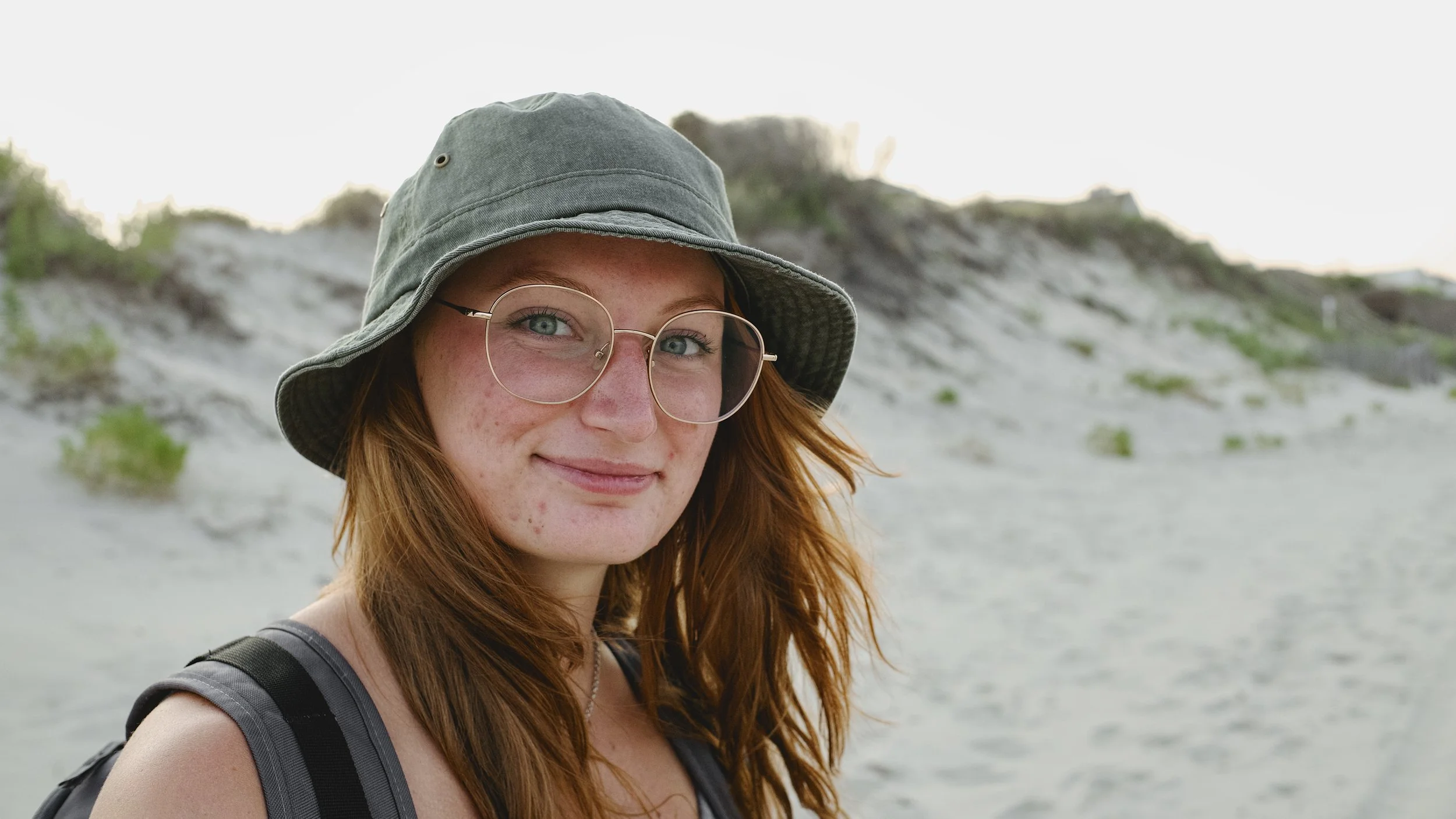 A young woman with red hair, glasses, and a green bucket hat smiling at the camera on a beach with dunes and greenery in the background.