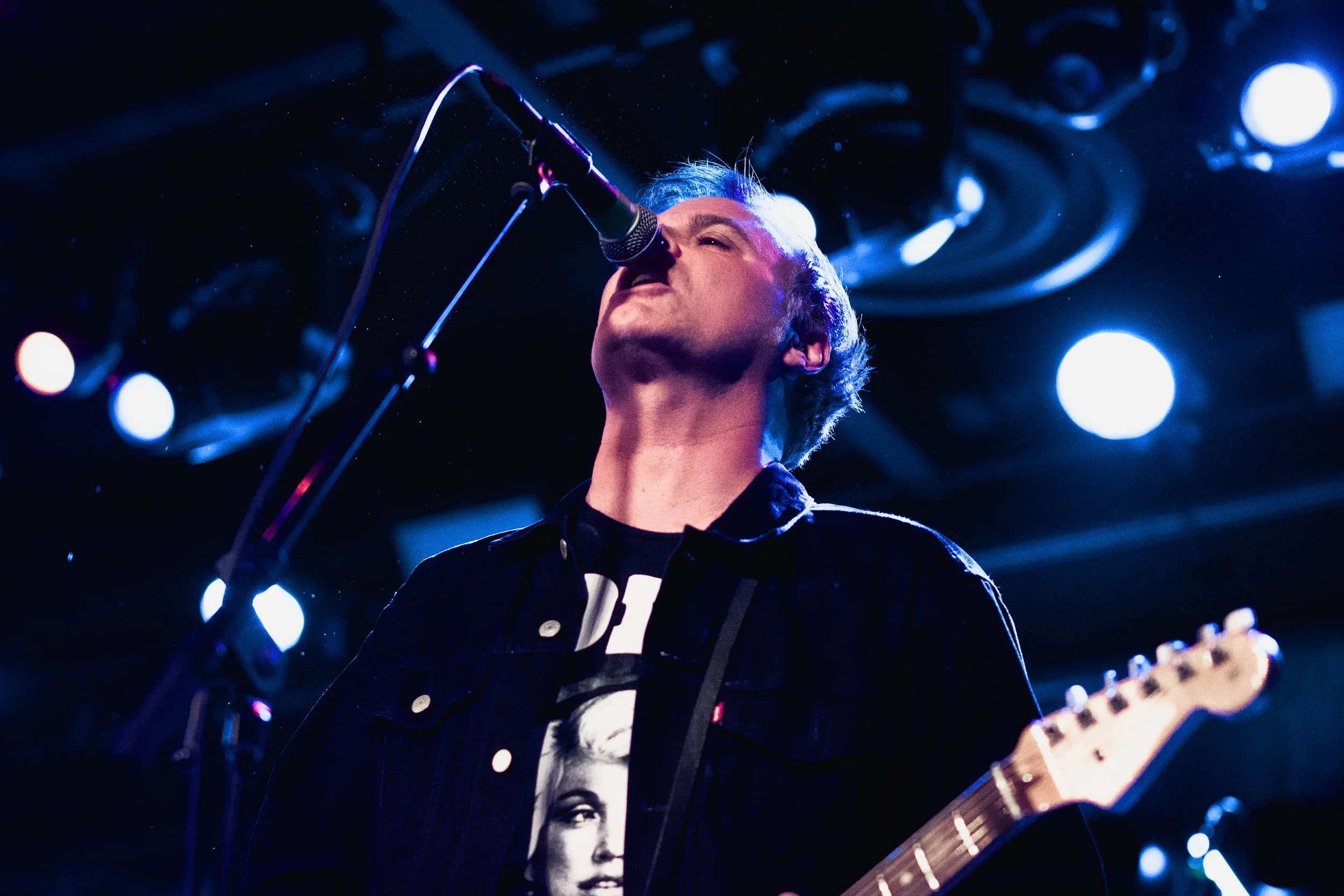Male musician singing into a microphone while playing an electric guitar during a live performance, with stage lighting in the background.