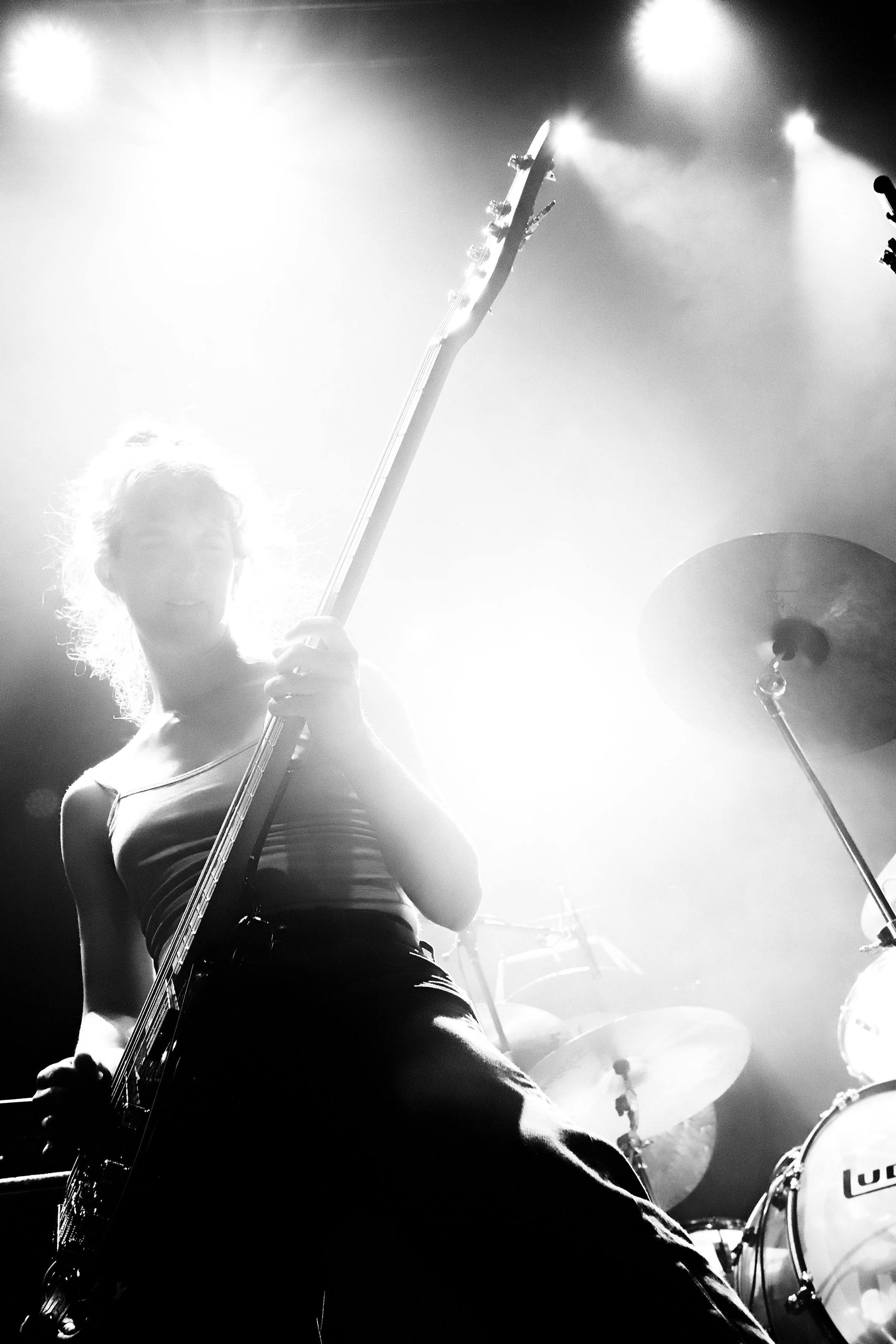 A woman playing an electric guitar on stage, backlit creating a silhouette with bright light shining from behind, with drums in the background.