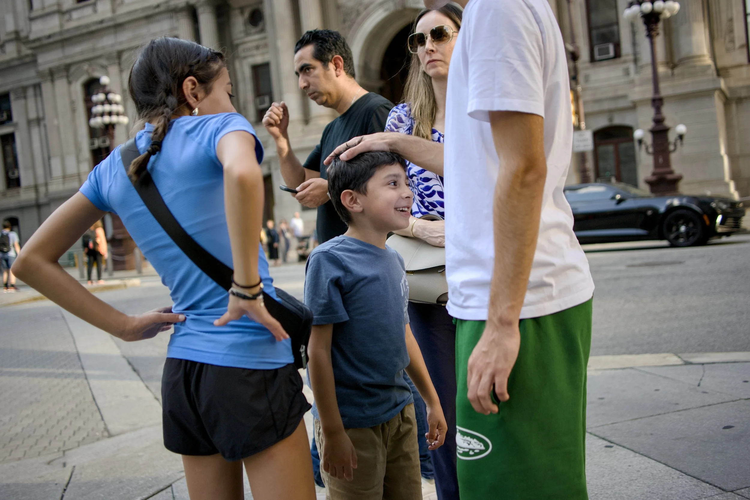 Group of people having a conversation on a city sidewalk, including a young girl in a blue shirt, a young boy smiling, a woman with sunglasses, and a man in a white shirt with green shorts, with historic buildings and cars in the background.