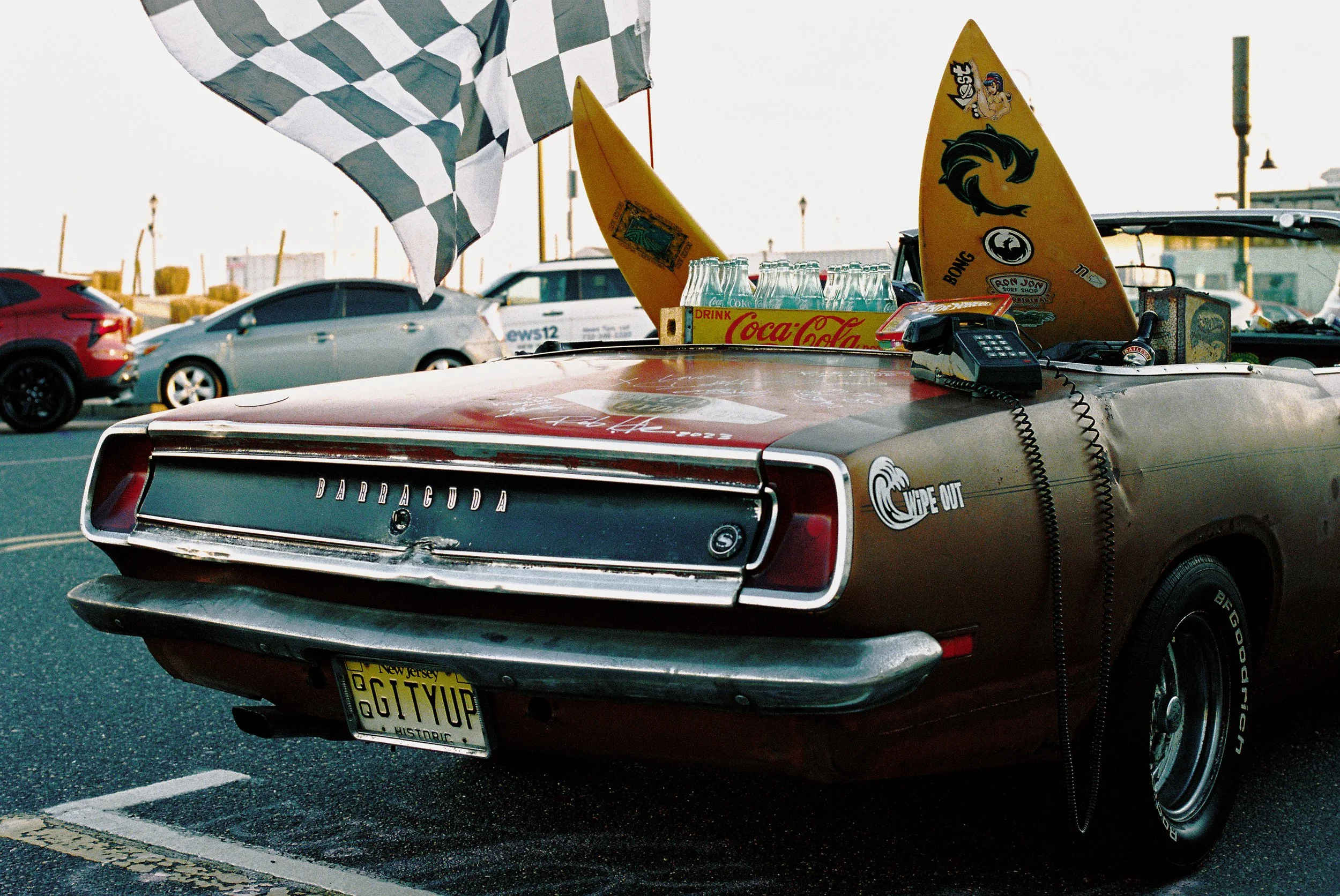 A vintage Barracuda car with a custom license plate reading "GGTYUP," parked in a lot with other vehicles in the background. The car's trunk is decorated with surfboards, a checkered flag, picnic items, a calculator, and stickers, including one that