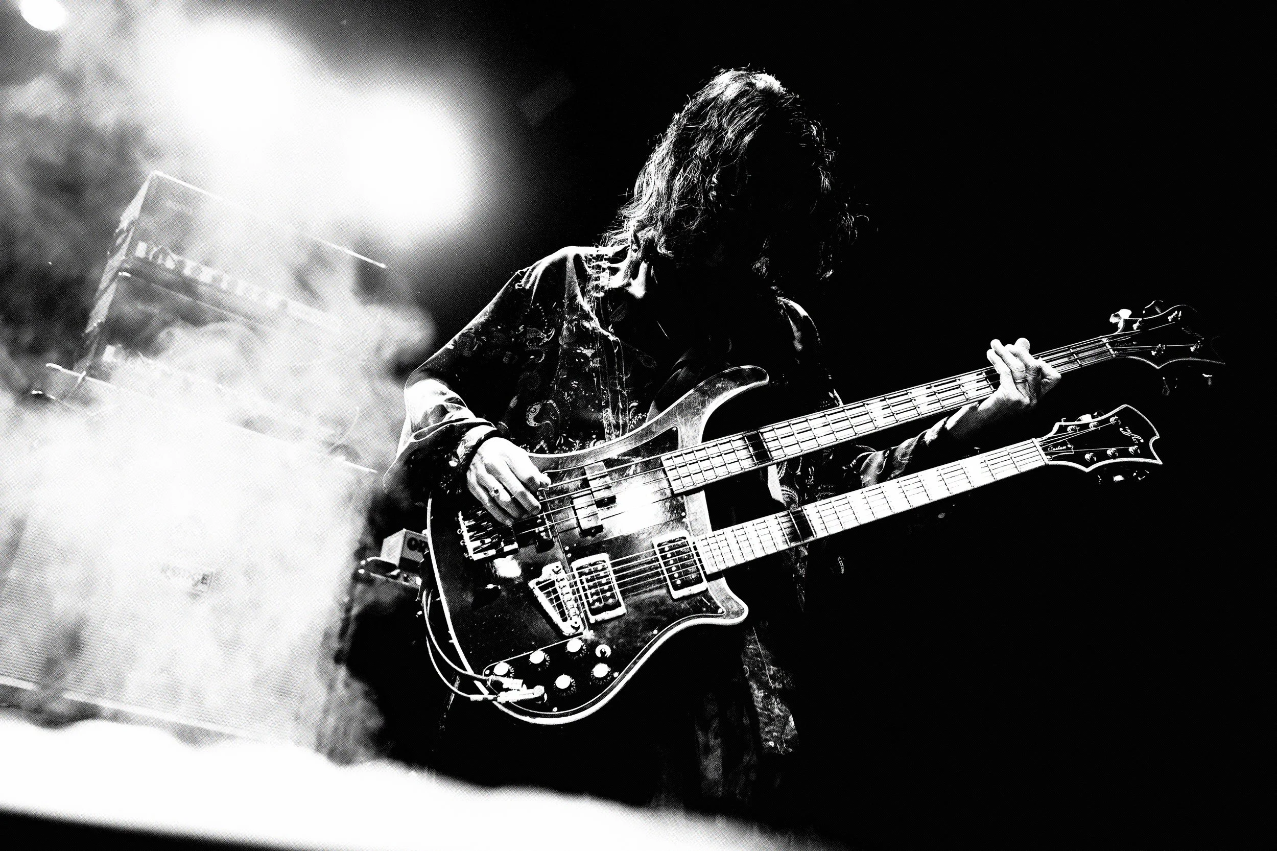 A musician playing eletric bass and guitar during a live concert, backlit with smoke and light effects, in black and white.
