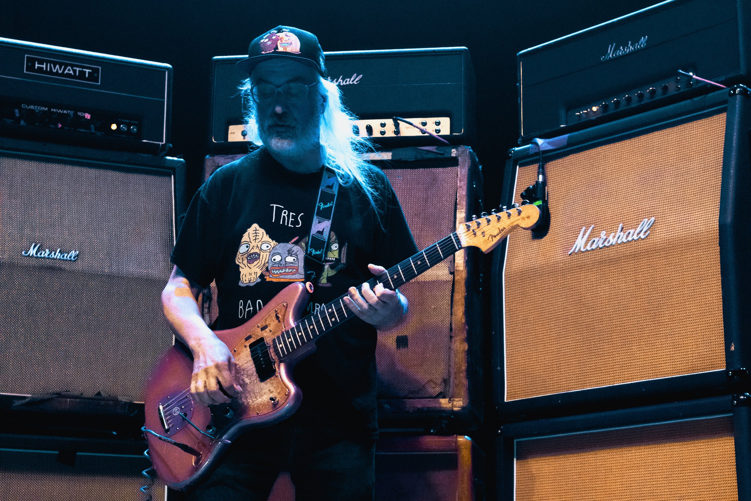 A man with long gray hair, glasses, and a beard playing an electric guitar on stage with a backdrop of Marshall and Hiwatt guitar amplifiers.