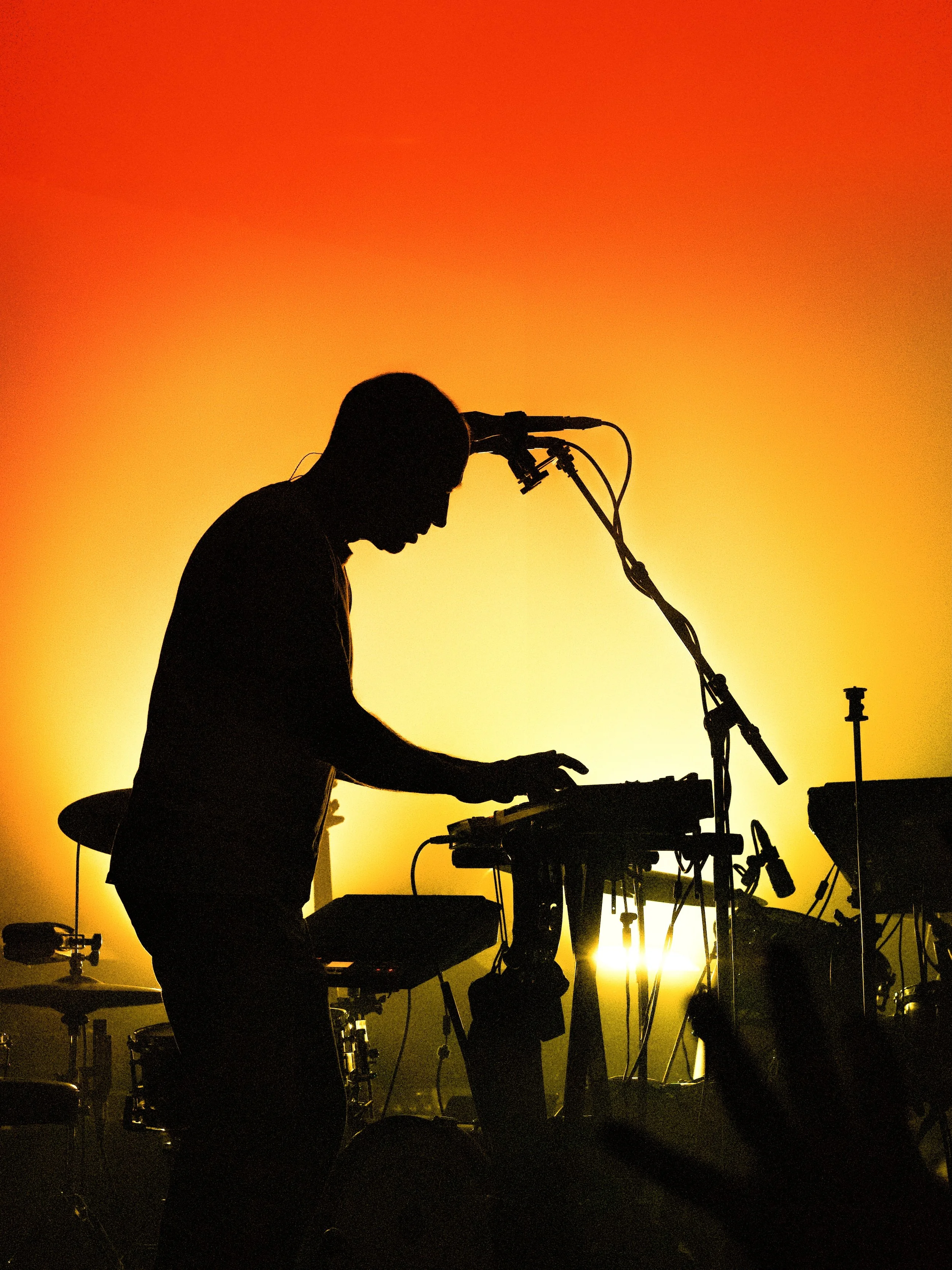 Silhouette of a musician playing electronic instruments on stage during a performance with a vibrant orange-yellow background.