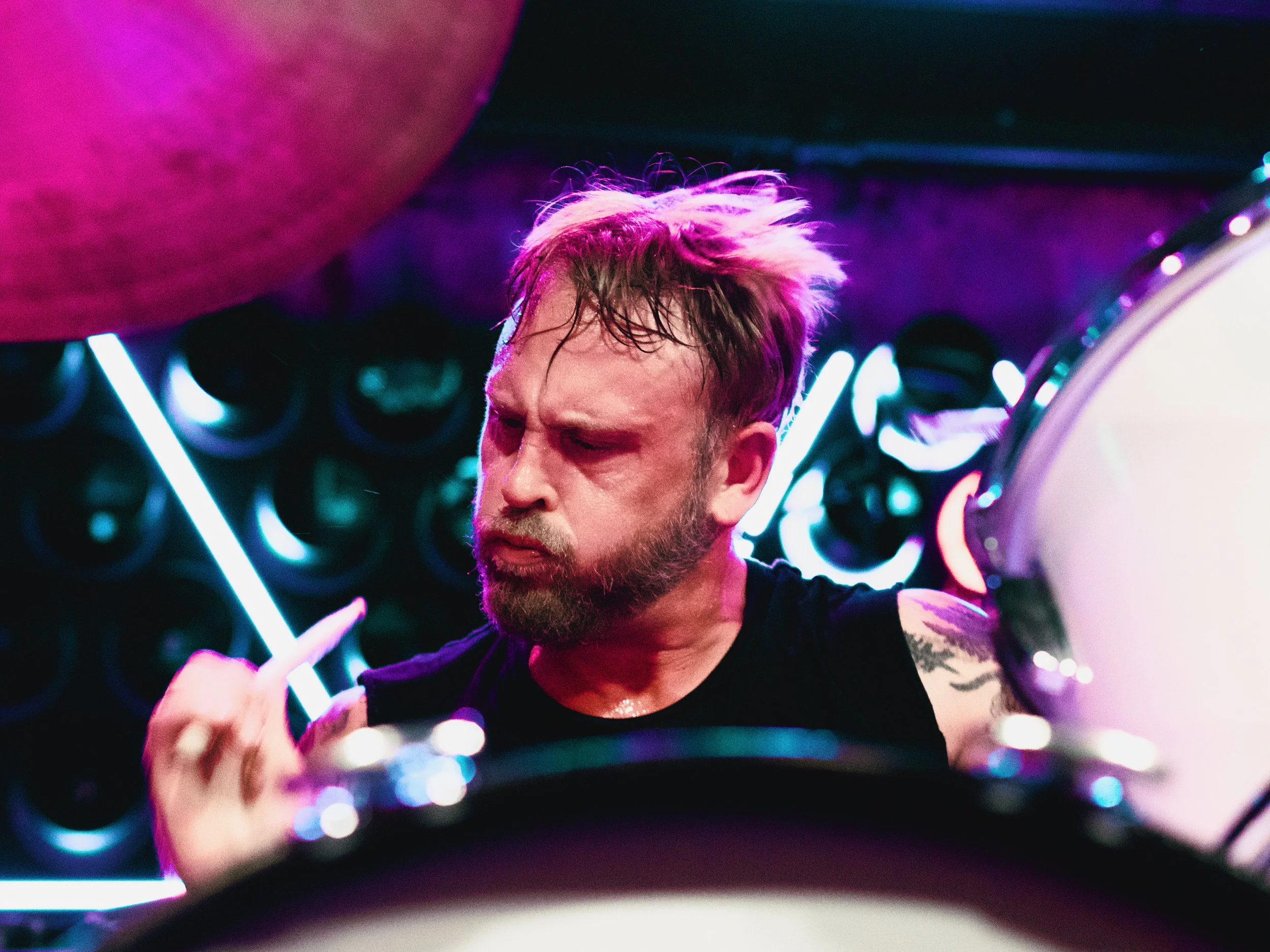 A man playing drums in a colorful, neon-lit setting, with intense focus on his face.