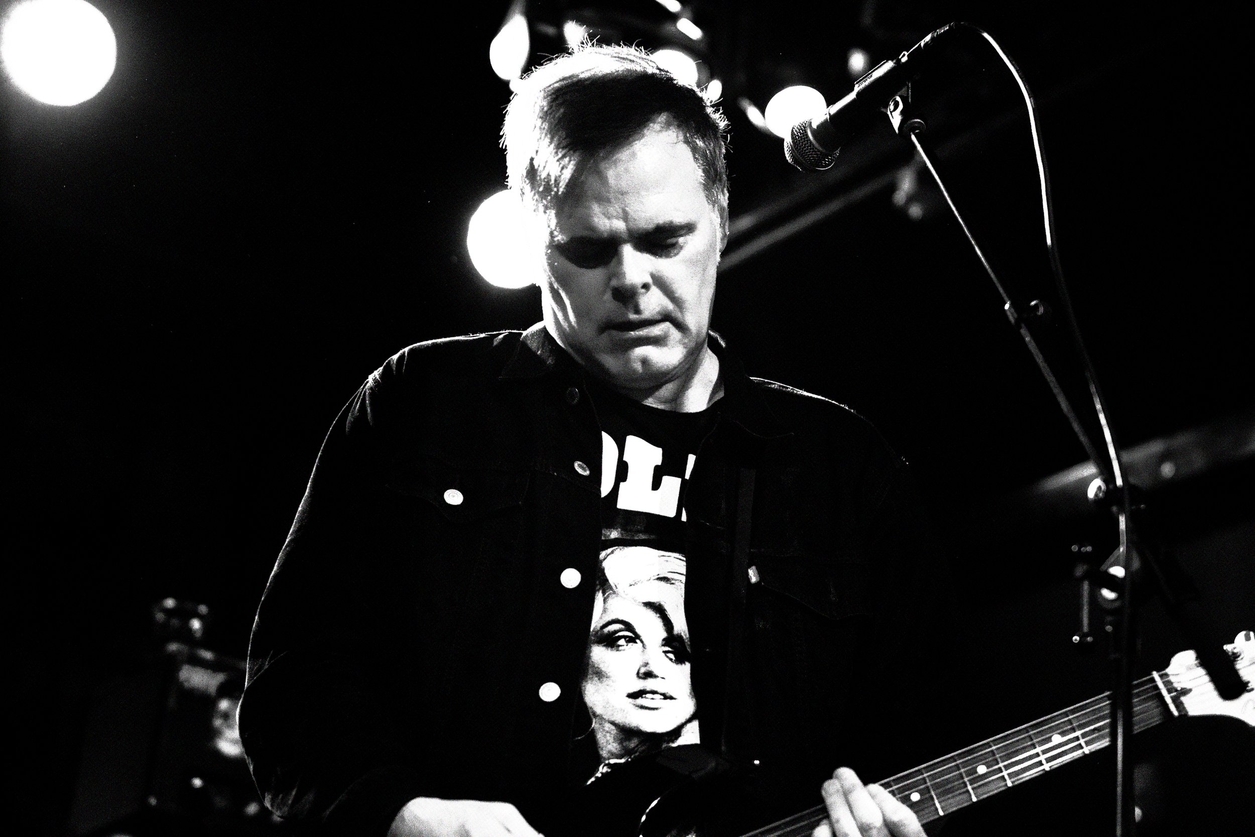 Black and white photo of a man playing an electric guitar during a live performance, with a microphone in front and stage lights in the background.