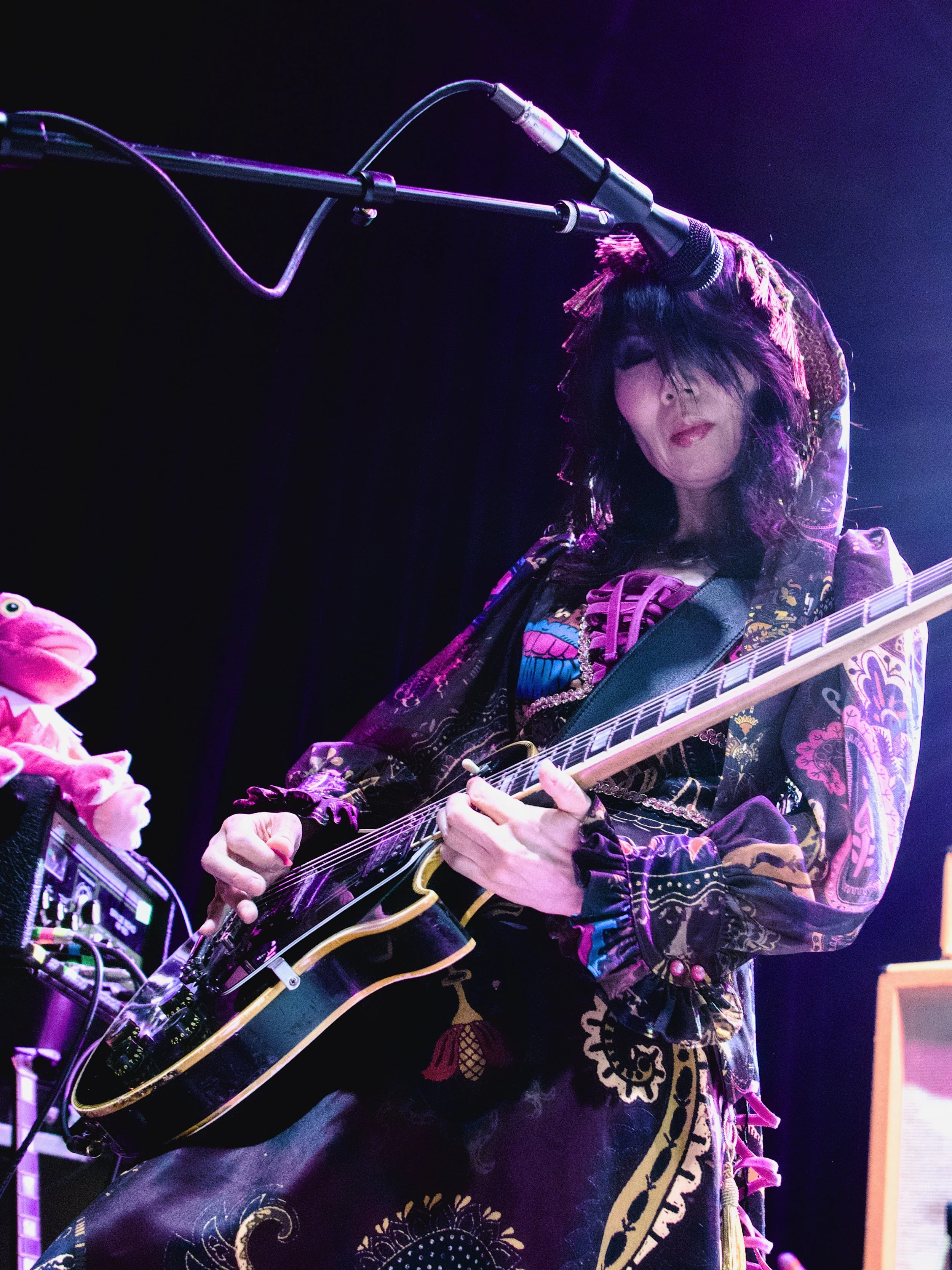 A woman performing on stage, playing an electric guitar with a microphone overhead, dressed in colorful, patterned clothing with a hood.