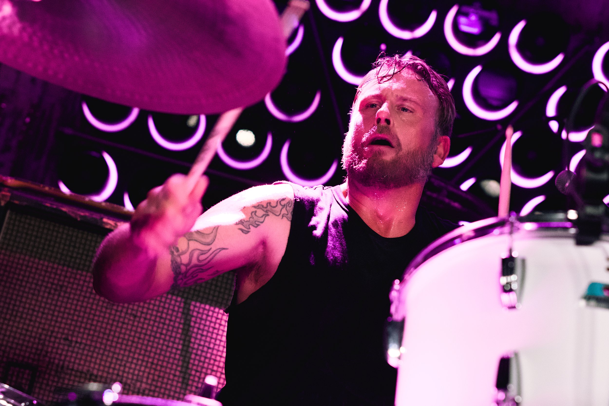 A man with a beard and tattoos on his arm playing drums during a live music performance, illuminated by purple and pink stage lights.