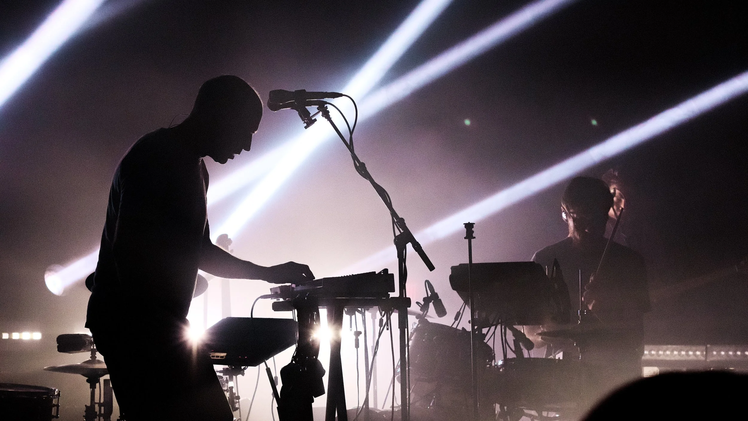 Silhouettes of two musicians performing on stage with bright stage lights and fog