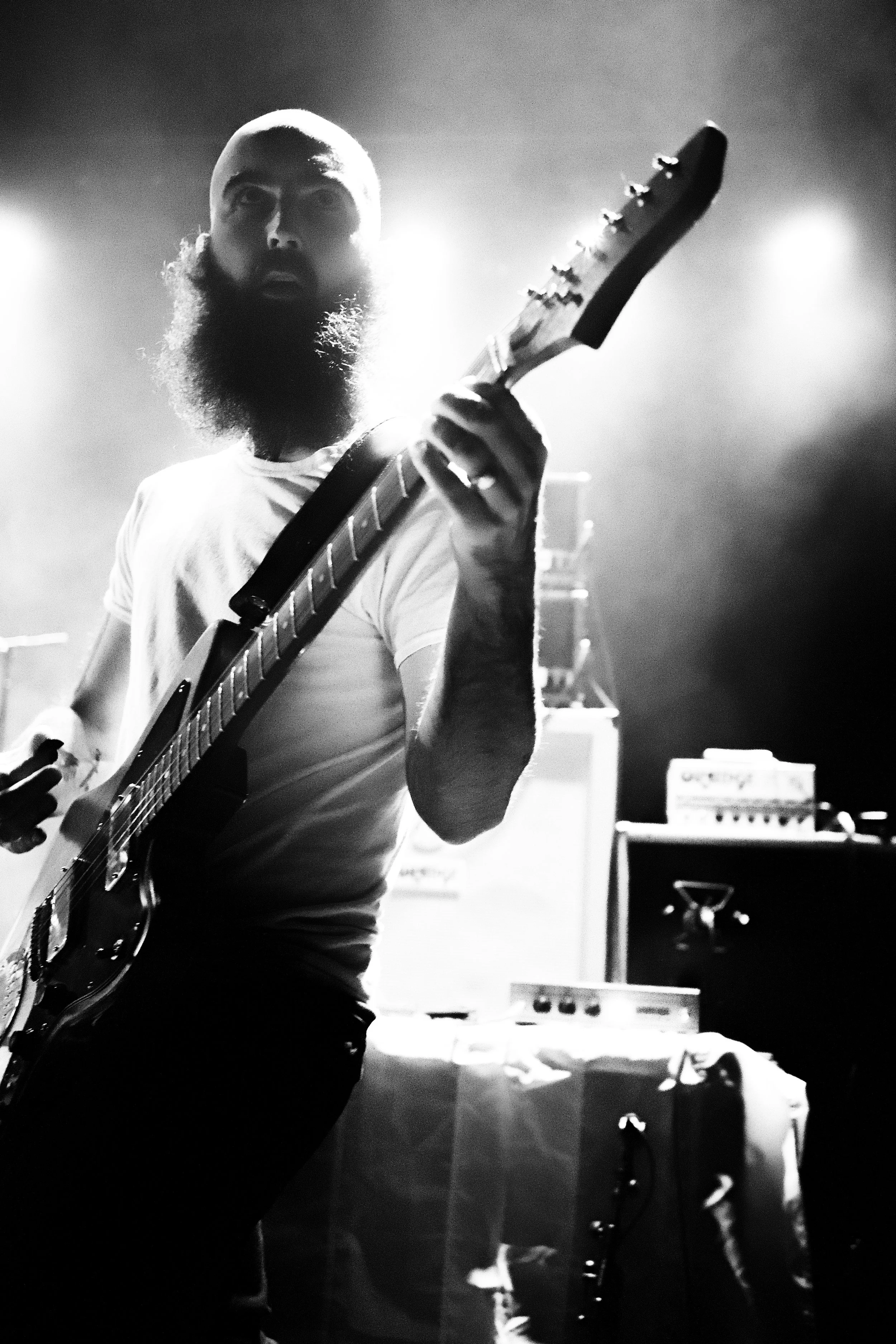 Black and white photo of a bearded man playing an electric guitar on stage, illuminated by stage lights.