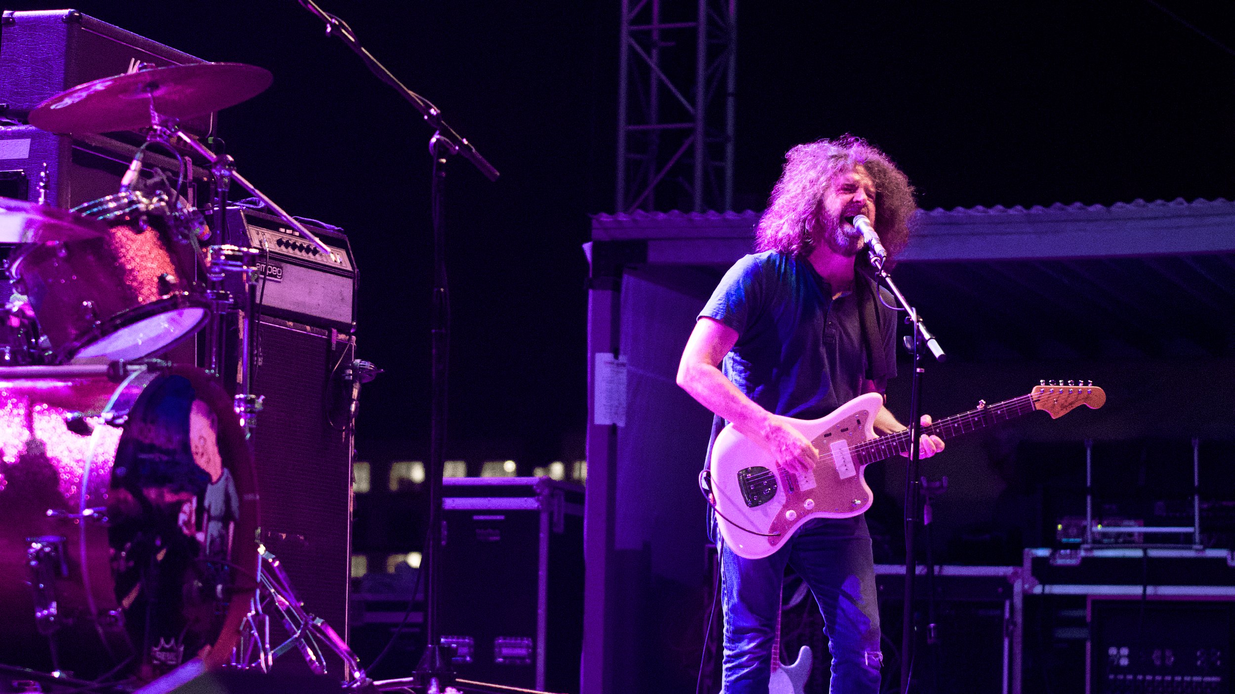 Musician with curly hair playing electric guitar and singing into a microphone on stage with purple lighting.