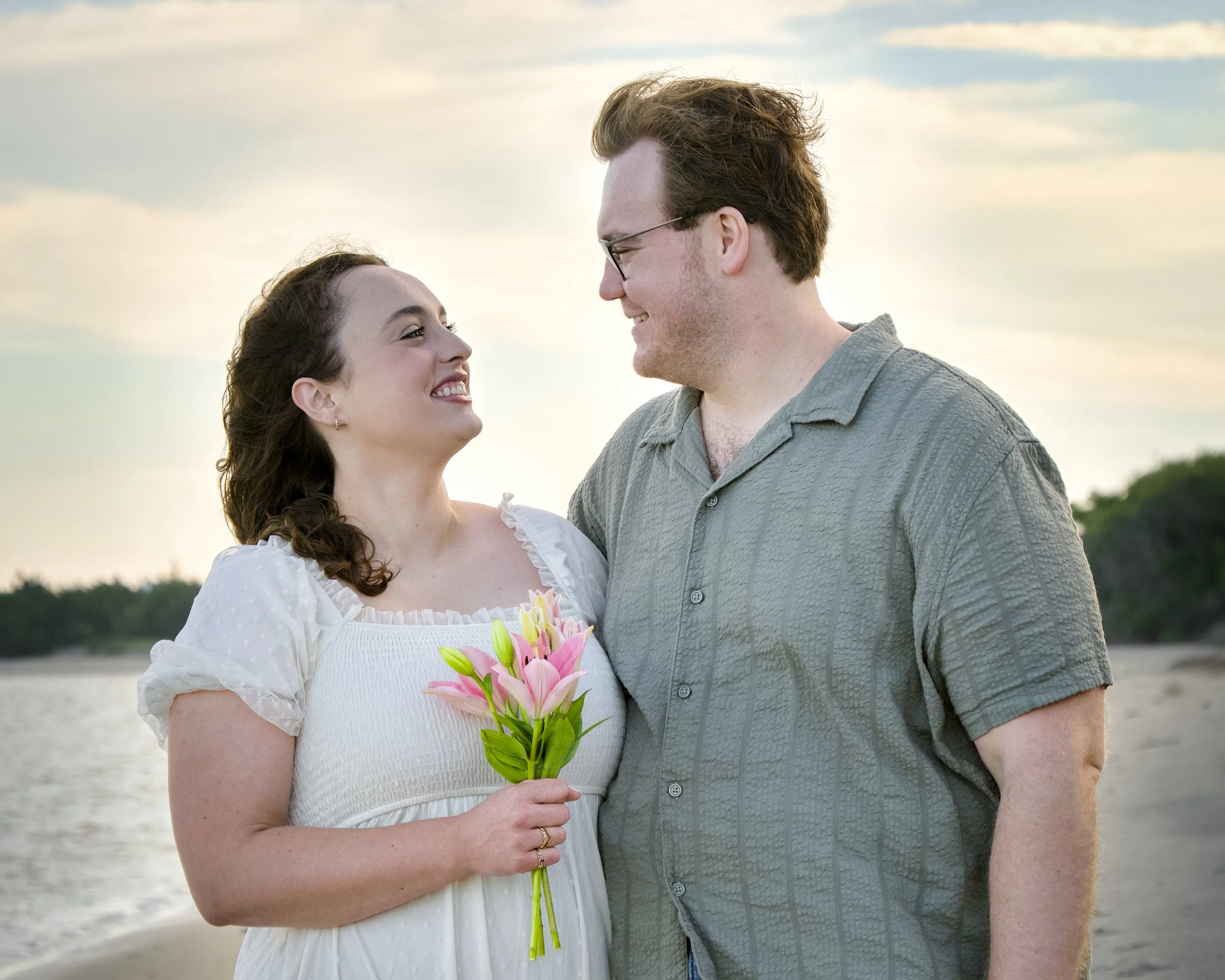 A young woman and a young man standing close together during sunset. The woman is holding a bouquet of pink lilies and smiling at the man. The man is smiling back and wearing glasses. The background features a calm body of water and a partly cloudy sky.