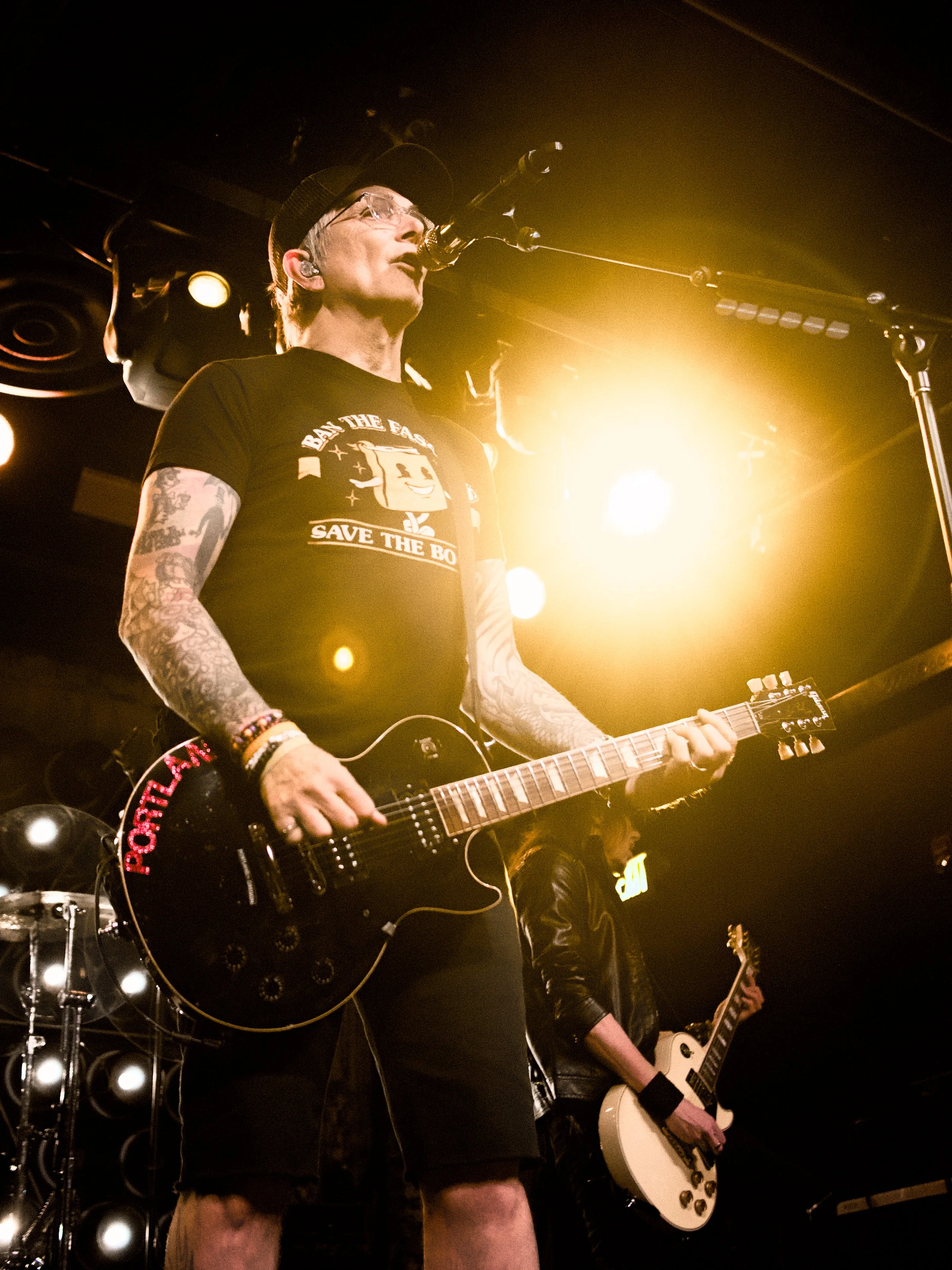 A musician with tattoos, wearing glasses, a cap, and a shirt that says 'Save the Bo,' is performing on stage with a guitar under bright stage lighting.