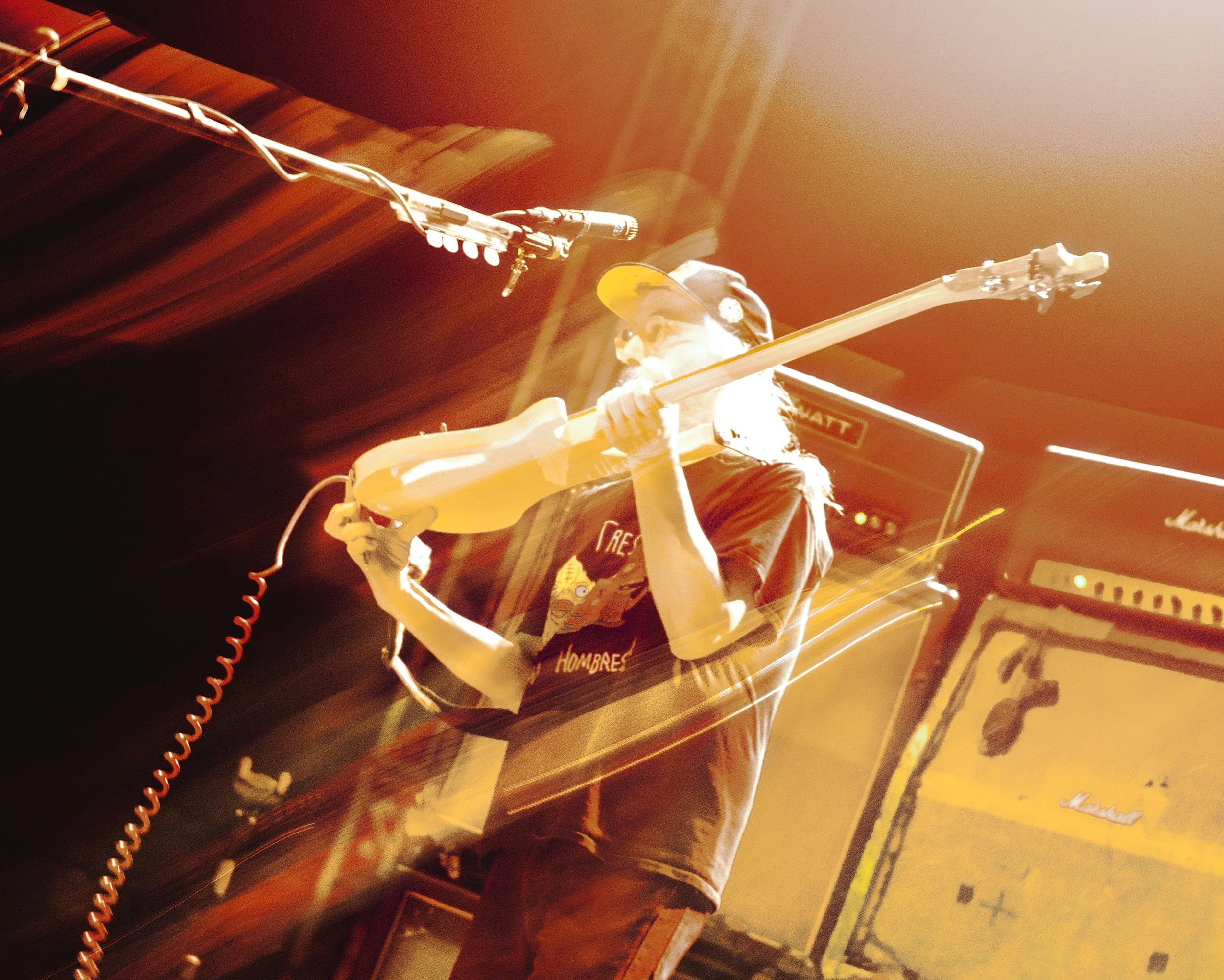 A person wearing a black cap and T-shirt playing an electric guitar on stage with large amplifiers behind them, in warm lighting.