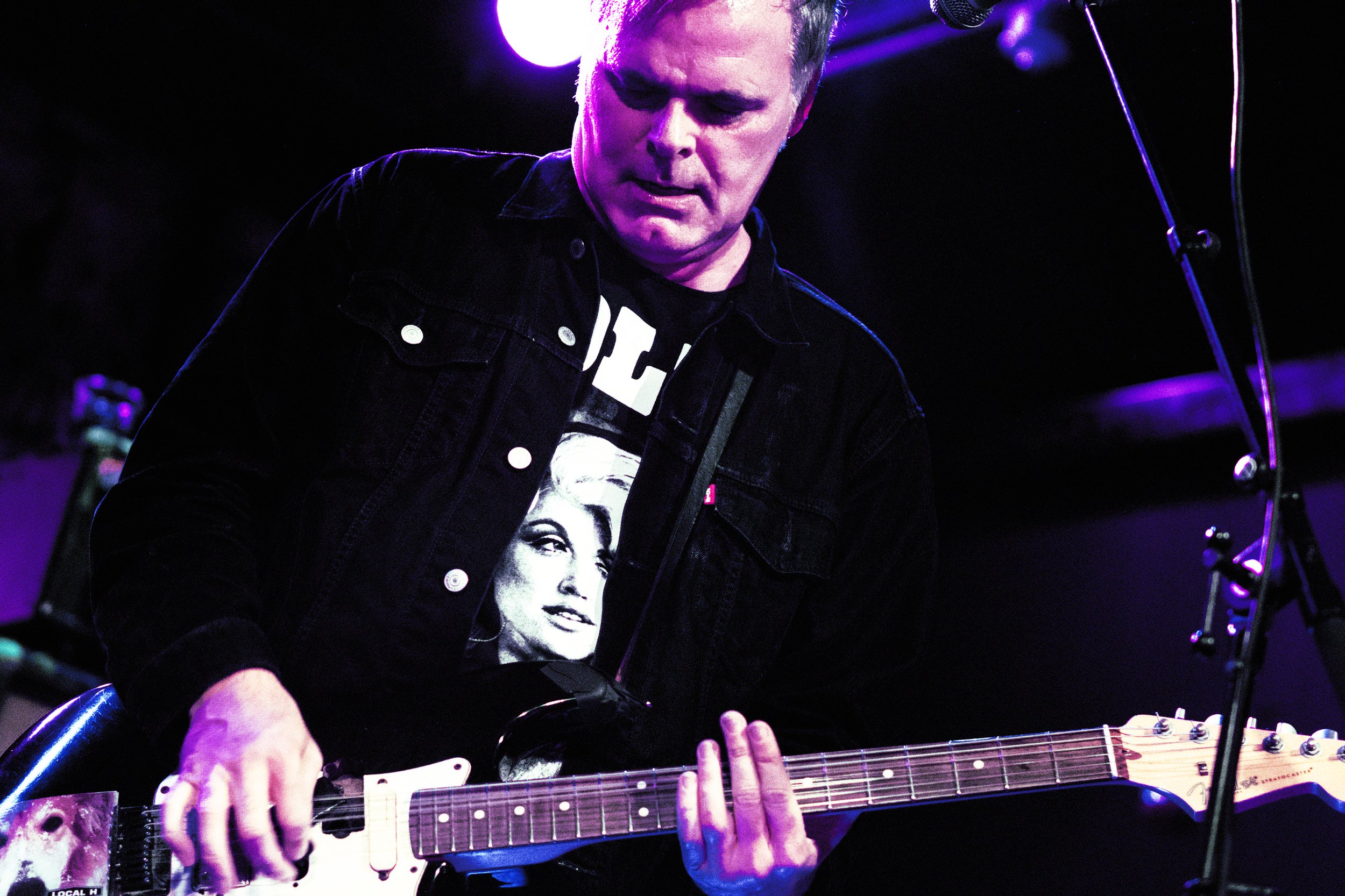 A man playing an electric guitar on stage, wearing a black jacket and a black T-shirt with a woman's face printed on it. The stage is lit with purple lights.