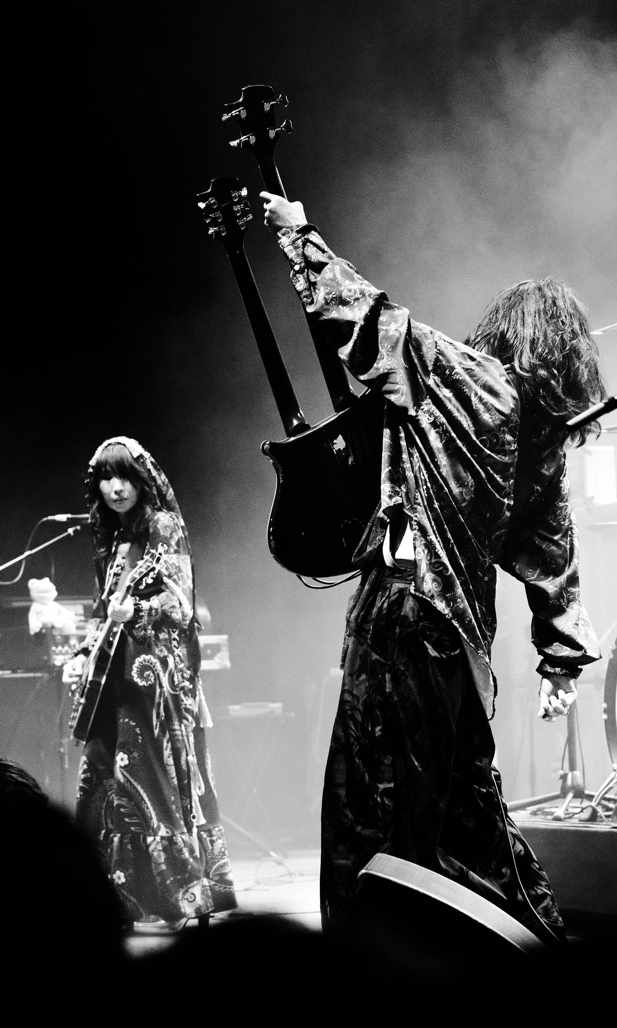 Black and white photo of two musicians on stage, one playing a guitar with their arm raised, the other playing a guitar and looking towards the camera, both wearing patterned, flowing clothing.