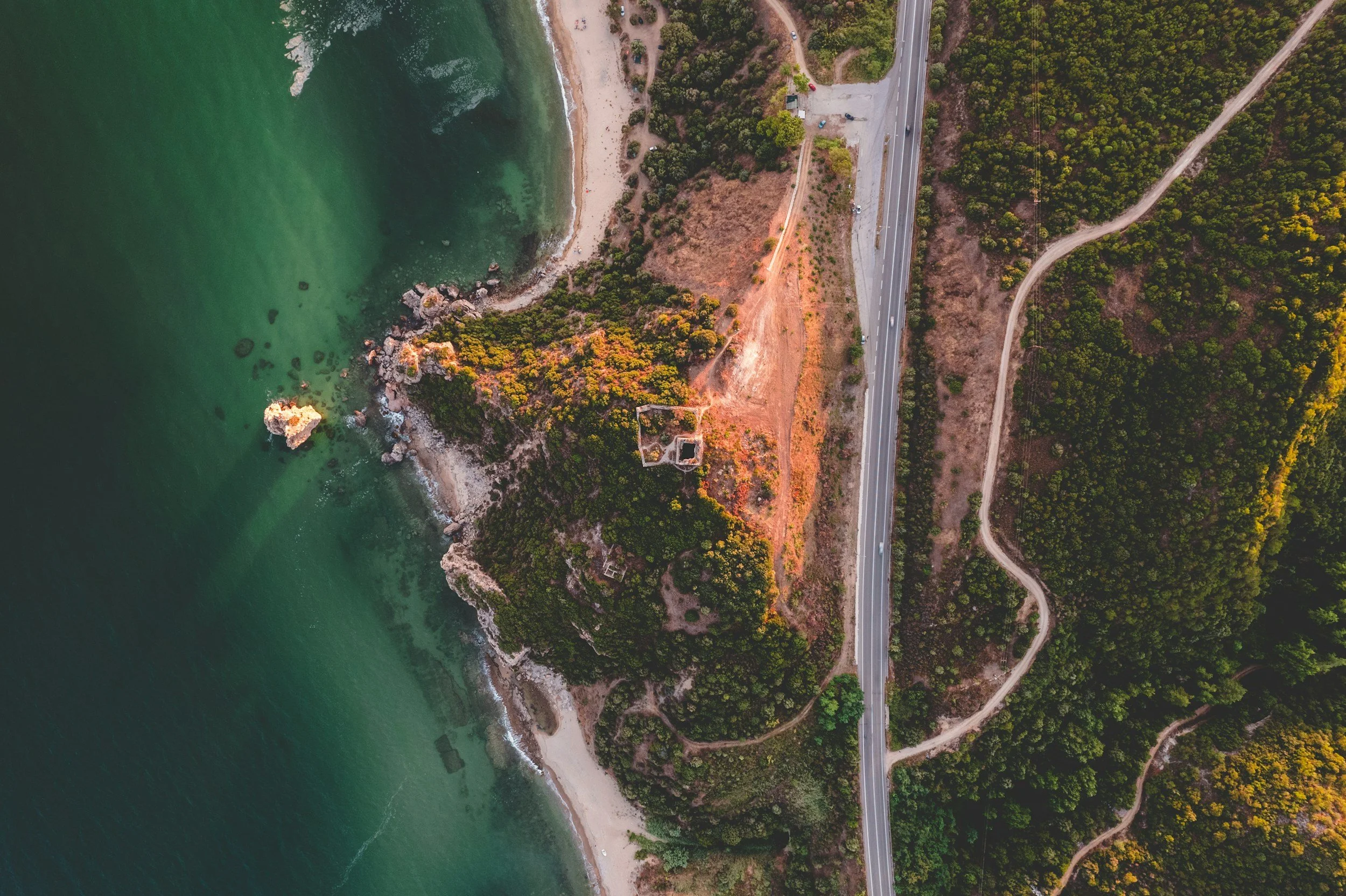 An aerial view of a coastal area showing a beach, green water, a rocky shoreline, a forested hill with some structures, and a highway running through the landscape with winding dirt paths nearby.