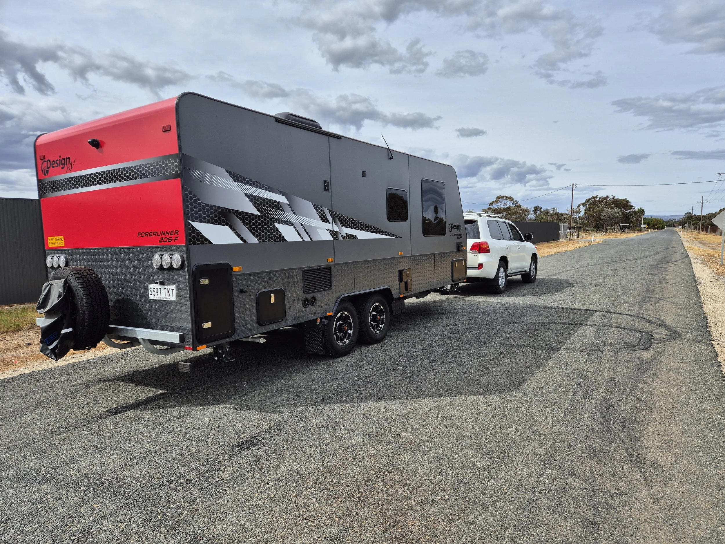A grey and red travel trailer with black accents parked on the side of an empty rural road with a white SUV attached behind it. The sky is partly cloudy.
