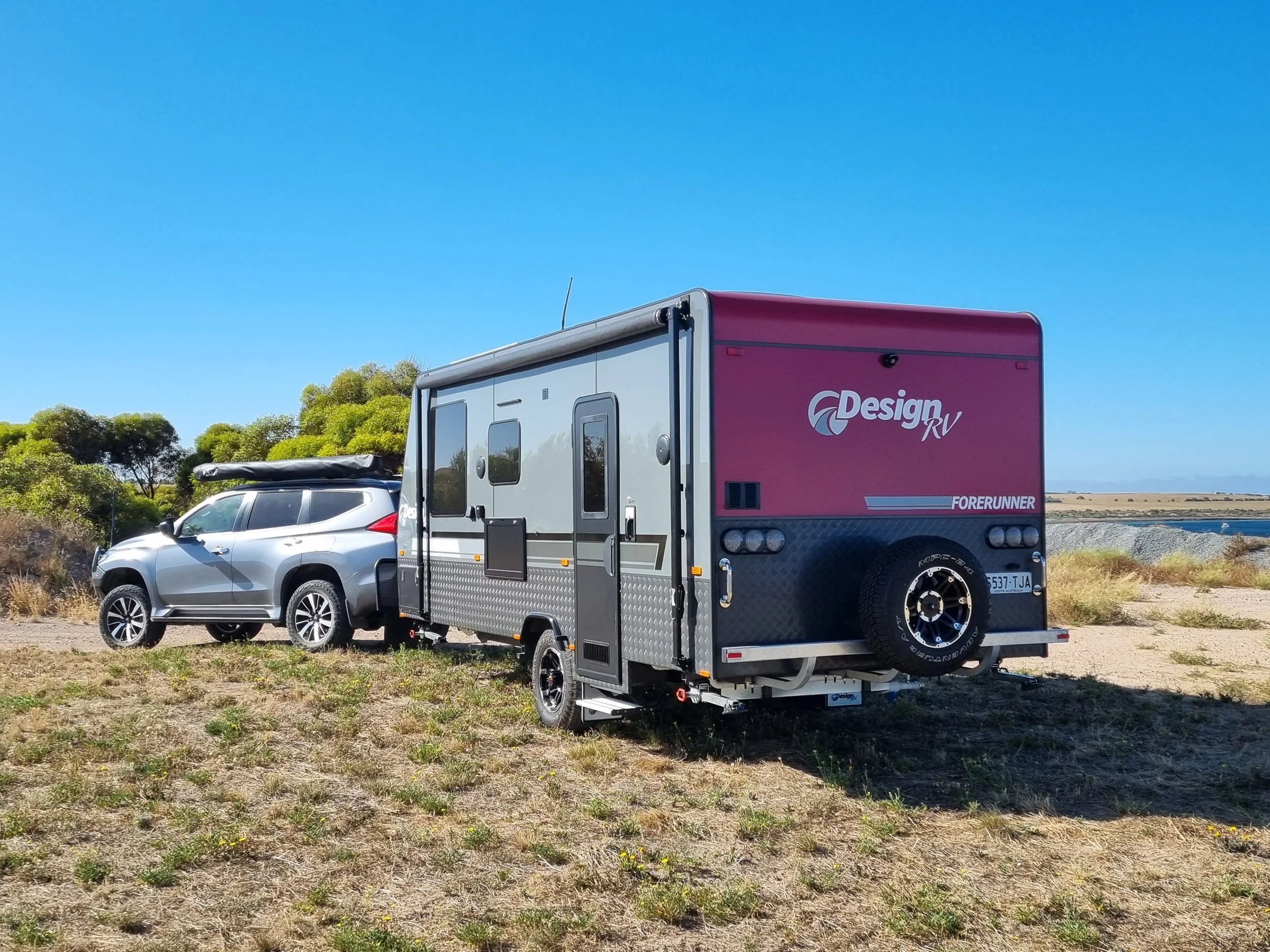 A silver suv pulling a maroon and gray design RV trailer with a spare tire at the back, parked on a grassy area near trees and a body of water under a clear blue sky.
