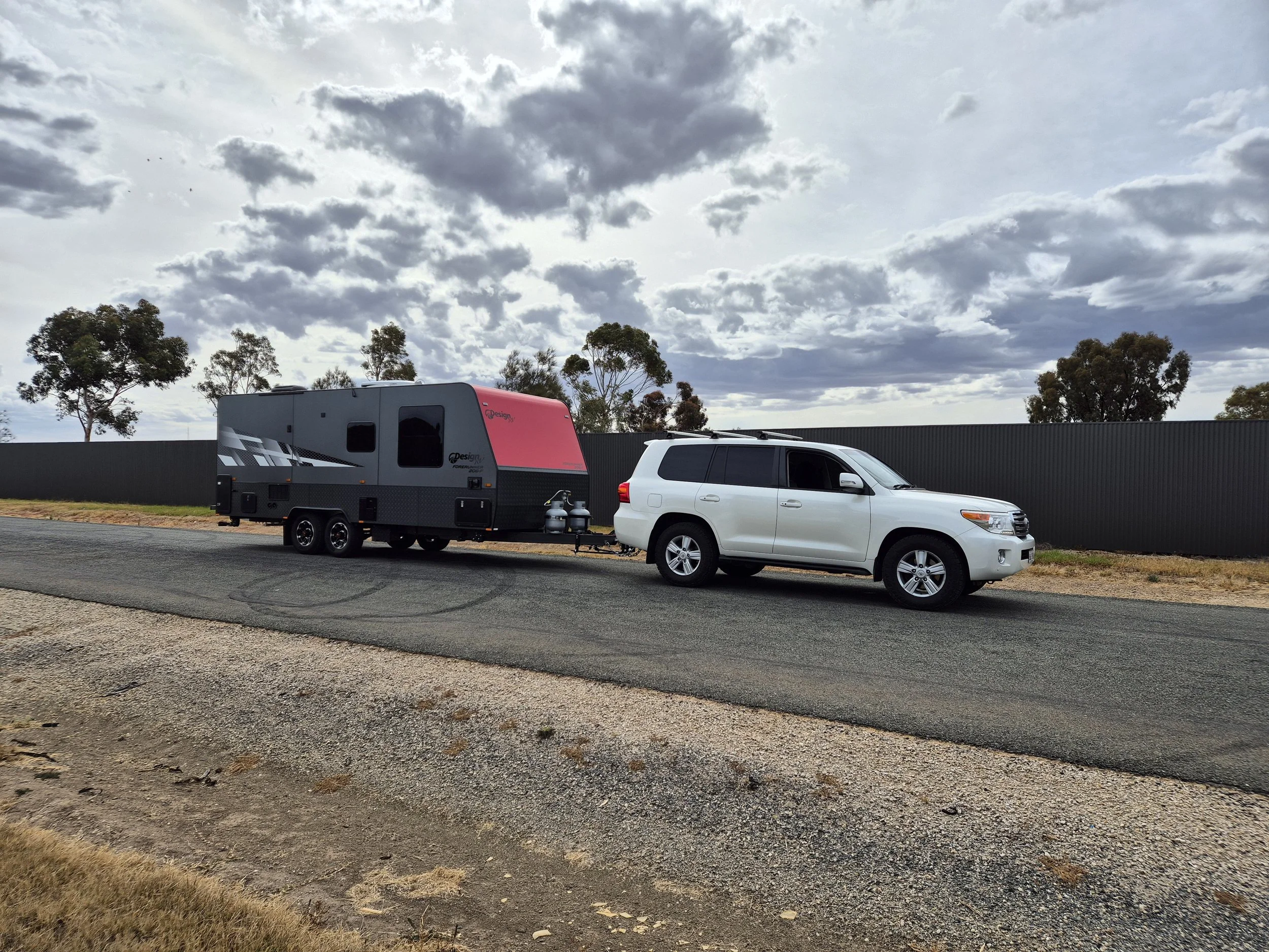 A white SUV parked on the side of the road attached to a black and pink travel trailer, under a partly cloudy sky, with trees and a black fence in the background.