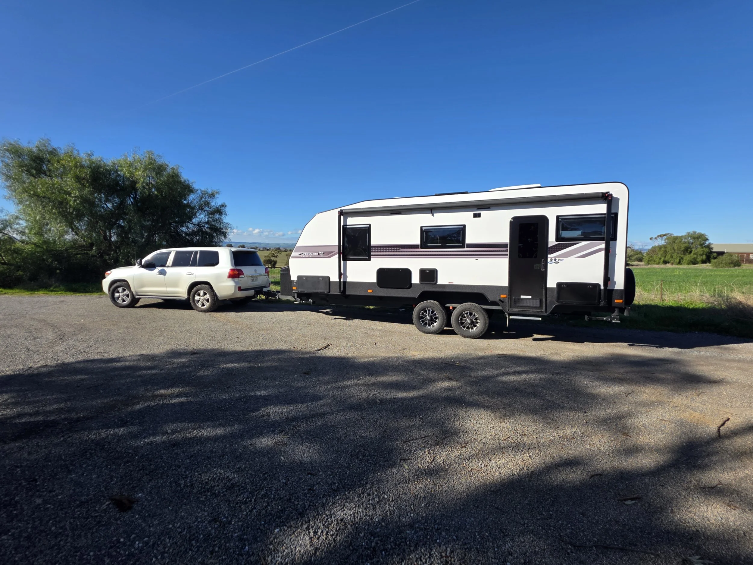 A white SUV parked on the side of the road, attached to a black-and-pink travel trailer. The scene features a cloudy sky with scattered clouds, a chain-link fence in the background, and some trees. The ground is a mix of gravel and asphalt.
